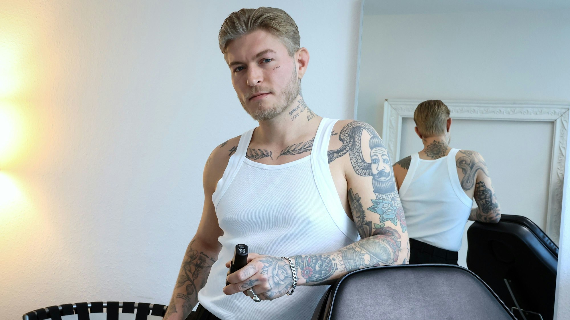 Das Bild zeigt Léon Sebastian Tiesler in einem weißen Unterhemd. Seine zahlreichen Tattoos auf dem Arm, am Brustbein, am Hals und im Gesicht sind gut zu erkennen.