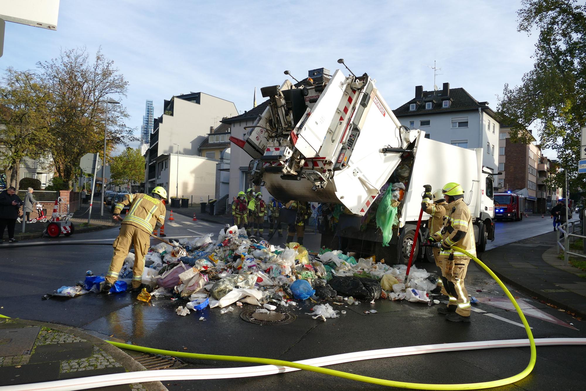 Feuerwehrleute löschen einen brennenden Müllwagen, der den Abfall auf die Straße entlädt.