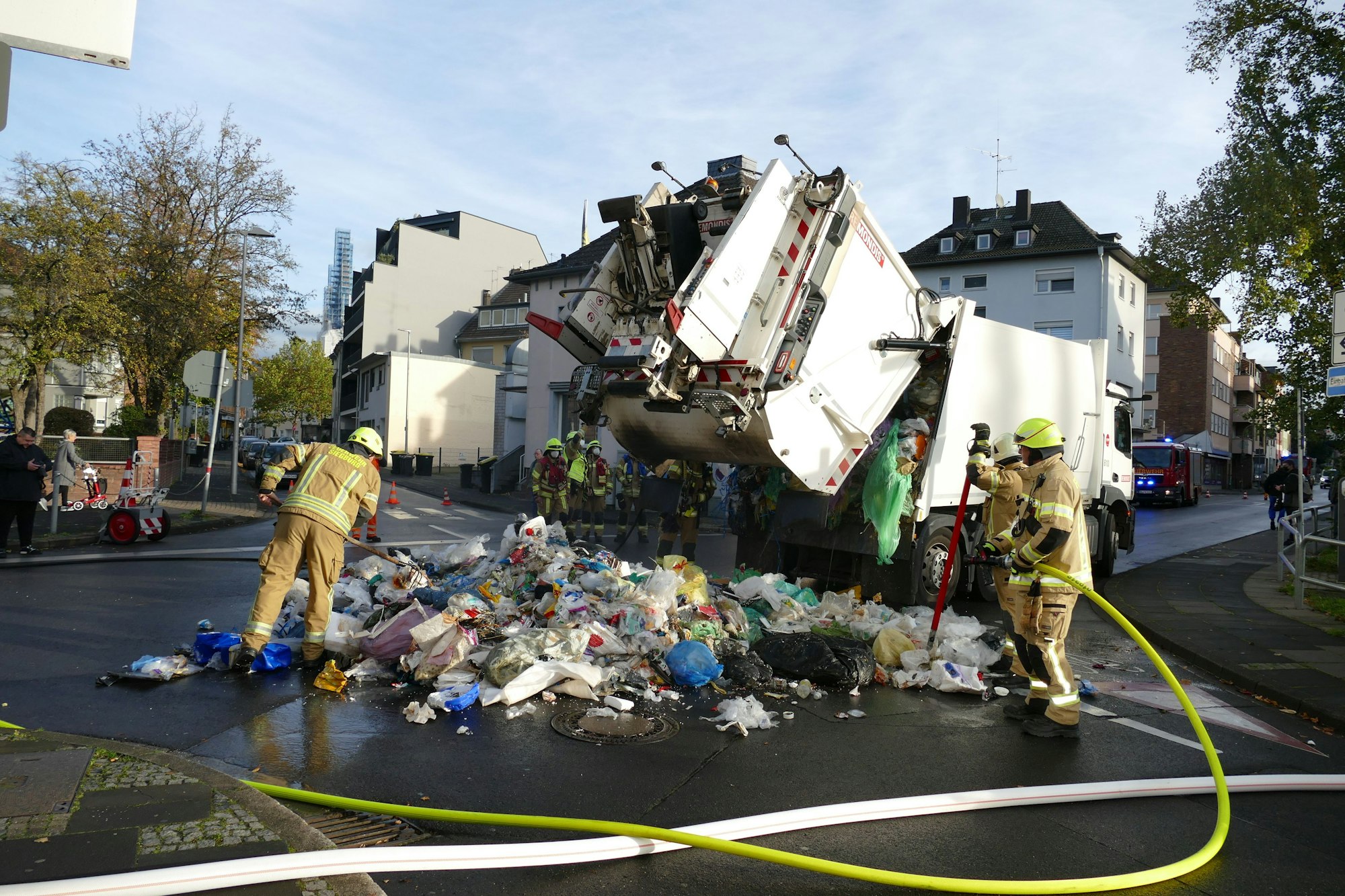 Feuerwehrleute löschen einen brennenden Müllwagen, der den Abfall auf die Straße entlädt.