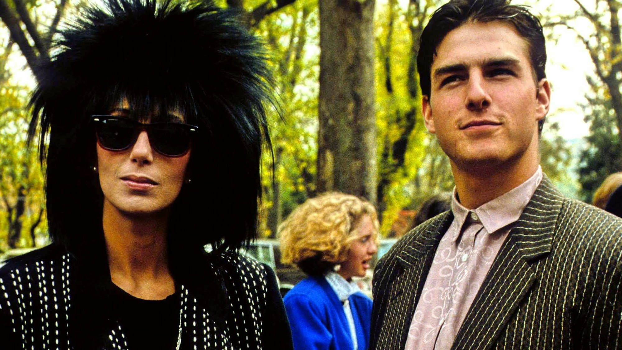 Cher mit schwarzer Fransenmähne und schwarzem Outfit, schwarzer Sonnenbrille. Neben ihr Tom Cruise, der leicht lächelt, mit braun-gestreiftem Jackett und altrosa Oberhemd. Beide stehen draußen, im Hintergrund sind Bäume zu sehen.