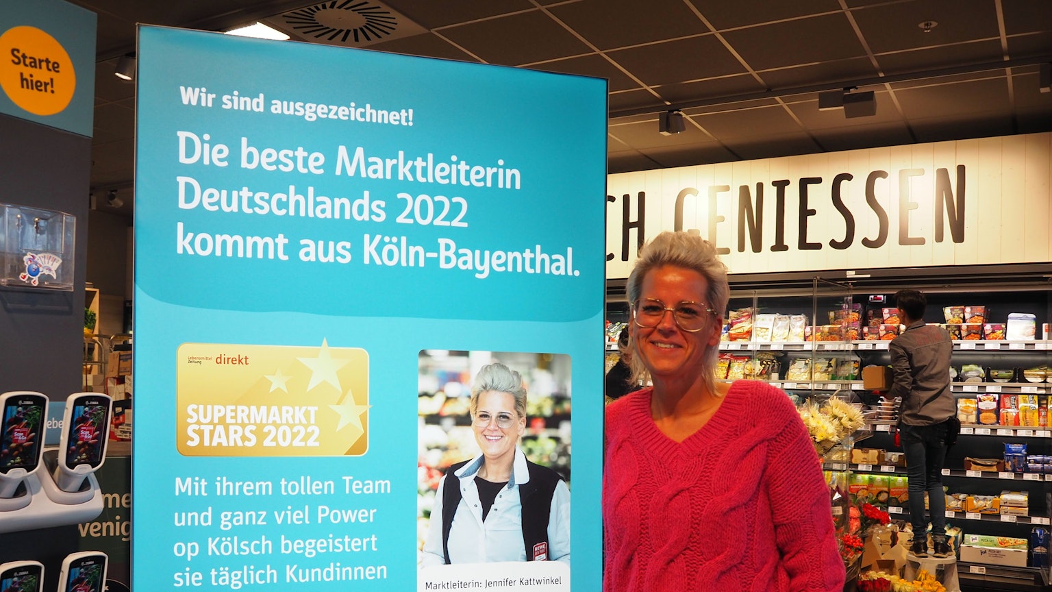 Jennifer Kattwinkel steht neben einem Porträt von sich selbst im Rewe-Markt in Köln-Bayenthal.