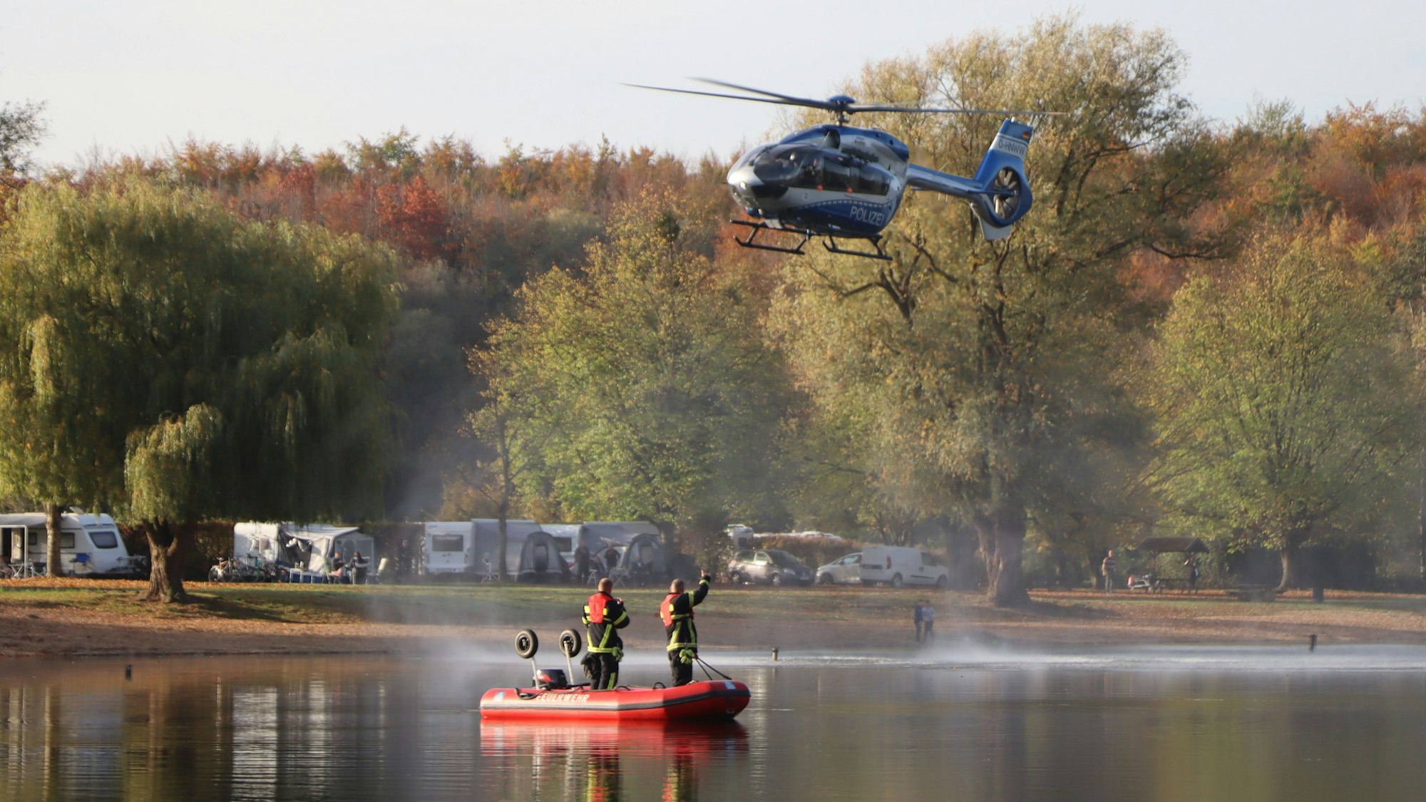 Feuerwehrleute in einem Rettungsboot auf einem See, über ihnen fliegt ein Hubschrauber.
