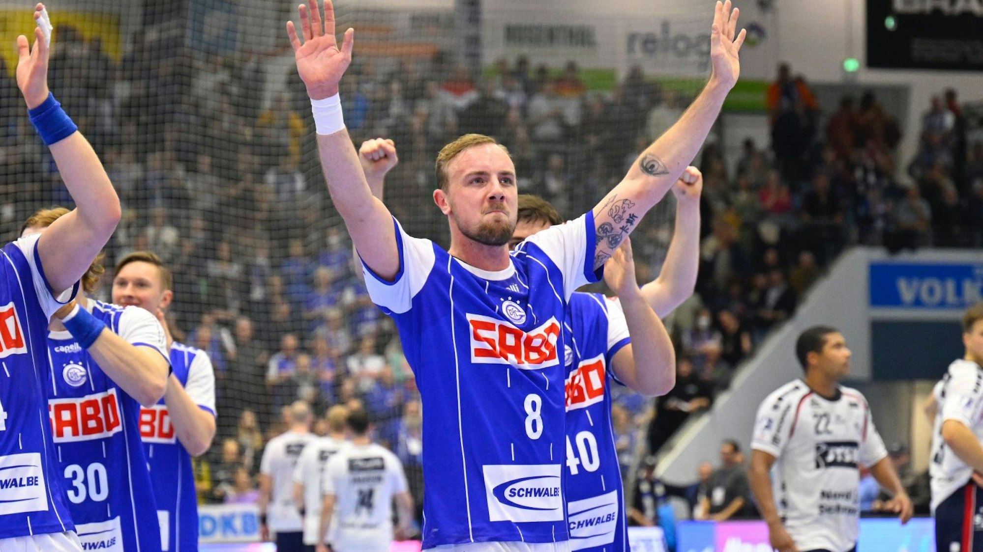 Handballspieler Lukas Blohme winkt den Zuschauern nach einem Spiel. Er trägt das blaue Trikot des VfL Gummersbach. Mannschaftskollegen stehen um ihn herum.