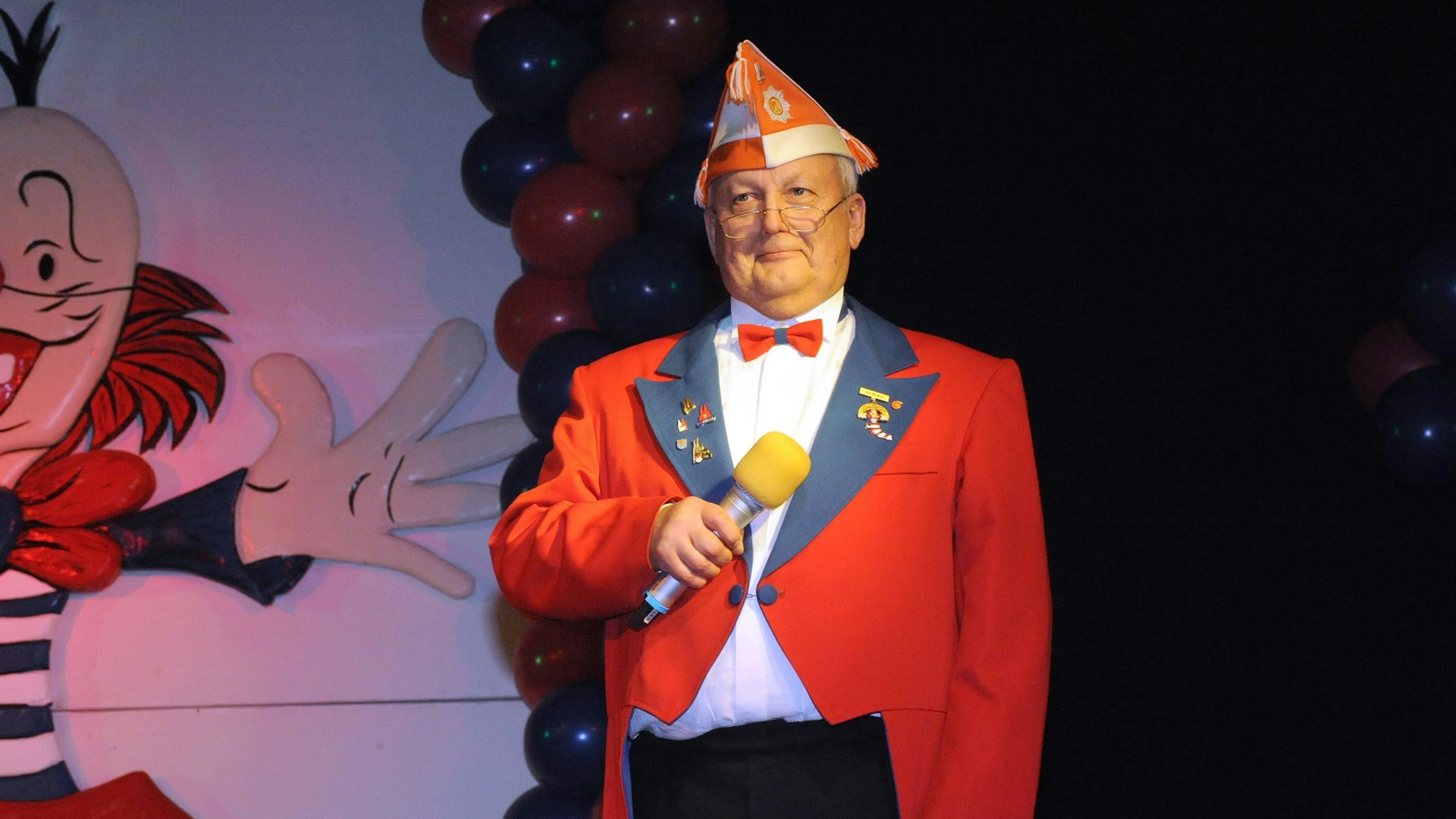 Winfried Reichwald im roten Frack der KG Kirchstraße mit Mikrofon auf der Sitzungsbühne.
