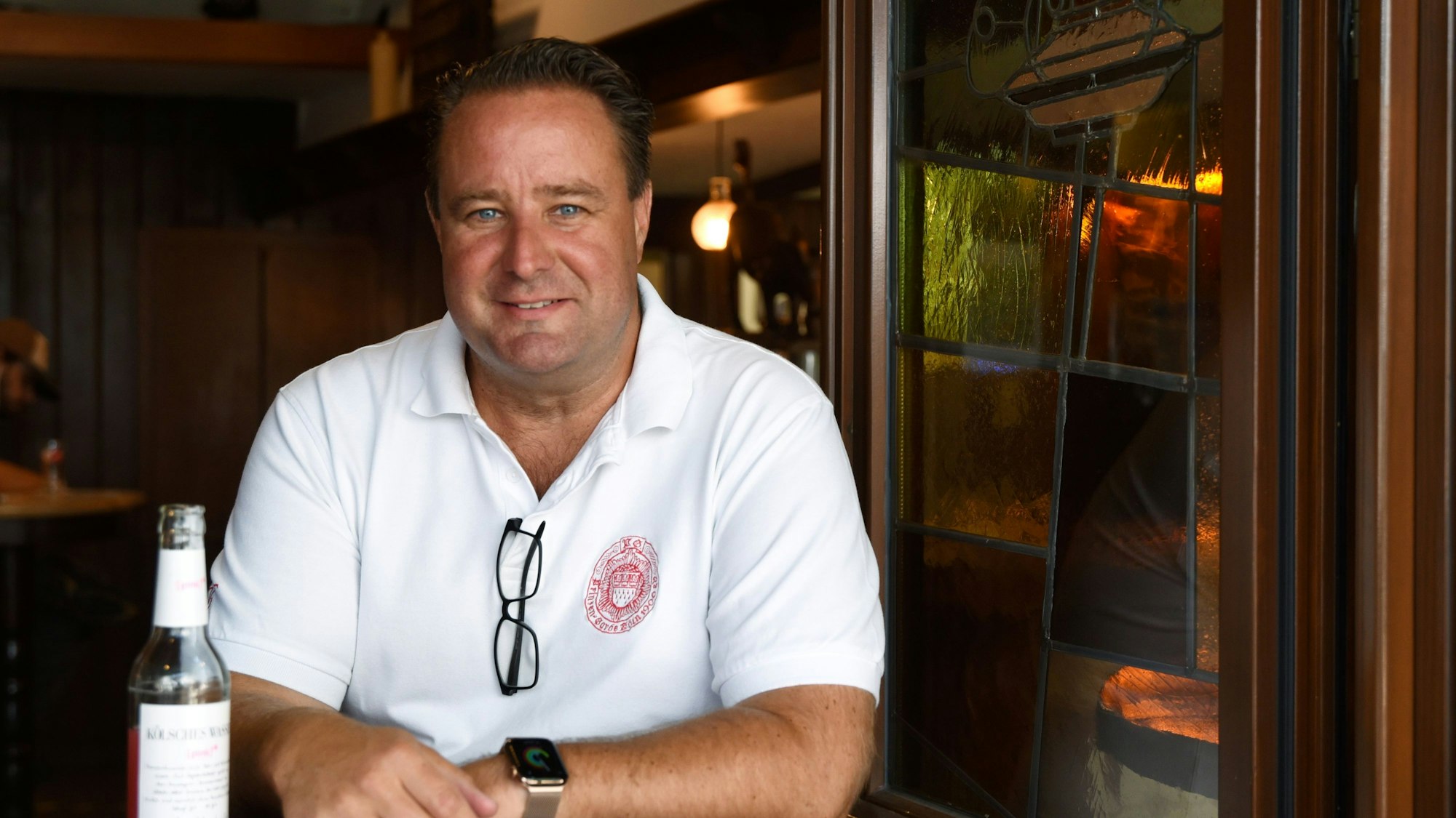 Alexander Manek steht in weißem Polohemd an einem Stehtisch einer seiner Gaststätten, vor ihm steht eine Limonade.