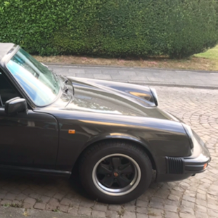 Der dunkel antrazit-farbene Porsche Targa Carrera steht auf der Straße vor einer grünen Hecke.