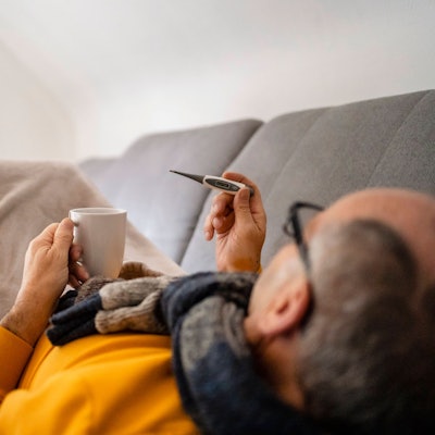 Eine Mann liegt auf der Couch und blickt auf ein Fieberthermometer.