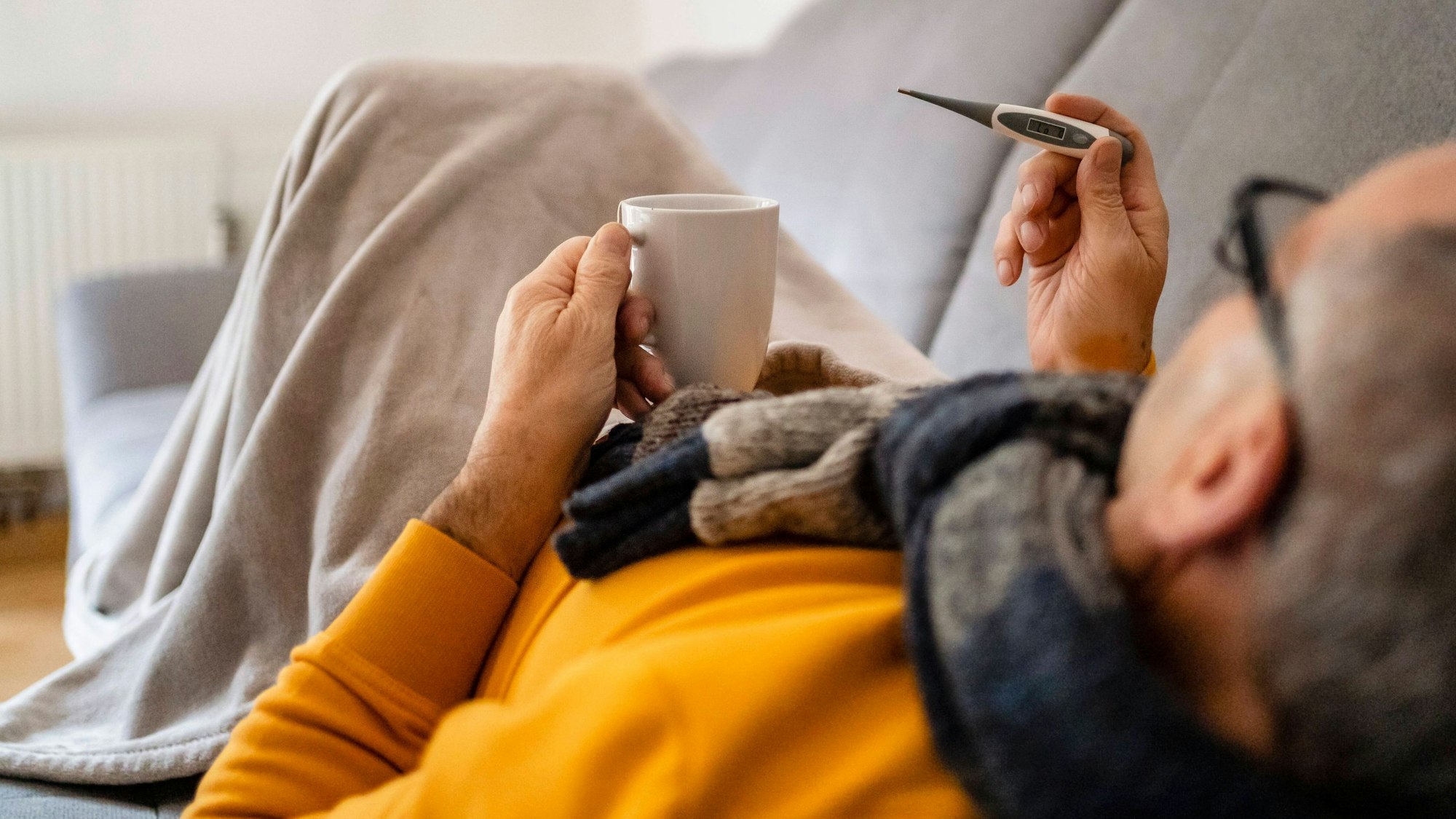 Eine Mann liegt auf der Couch und blickt auf ein Fieberthermometer.