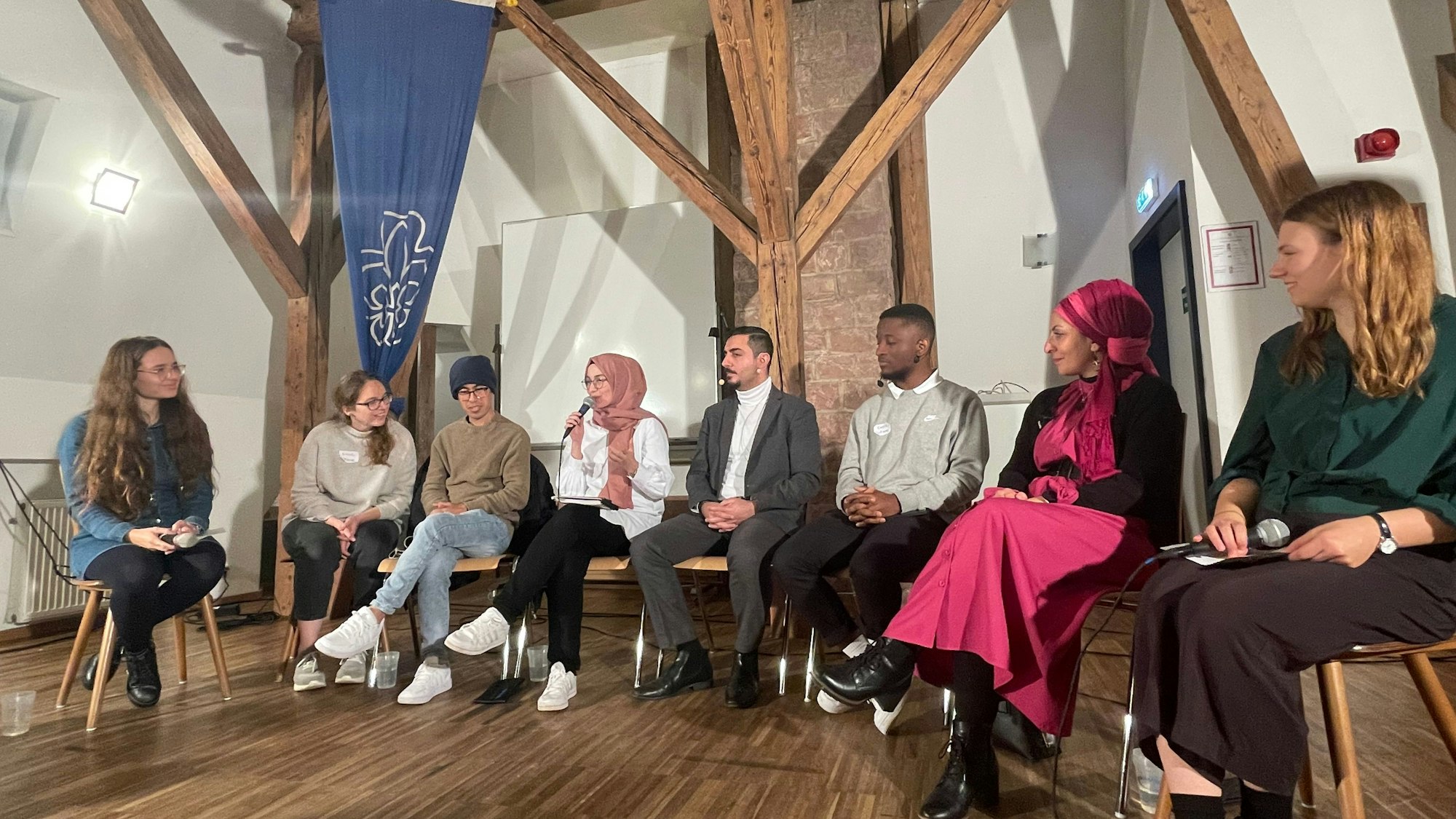 Die Mitgliederinnen und Mitglieder des Vereins Coexister sitzen in einem Stuhlkreis zusammen. Ein Mädchen in der Mitte hält mit einem Mikrofon in der Hand eine Rede, die anderen Anwesenden hören ihr zu.