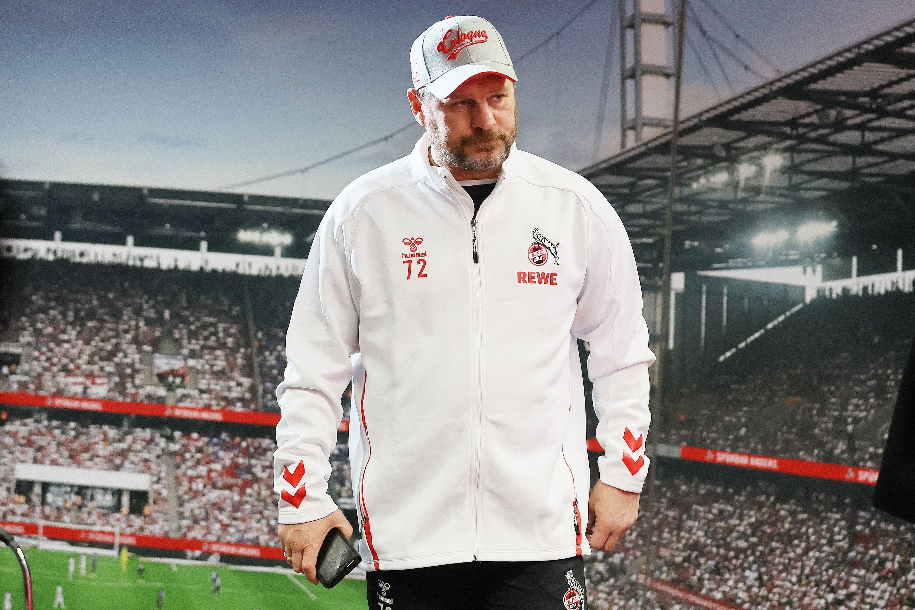 Steffen Baumgart steht im Pressesaal des Rhein-Energie-Stadions vor einem Bild der vollbesetzten Ränge. Der FC-Trainer trägt eine weiße Trainingsjacke und Kappe und schaut etwas morgenmuffelig.