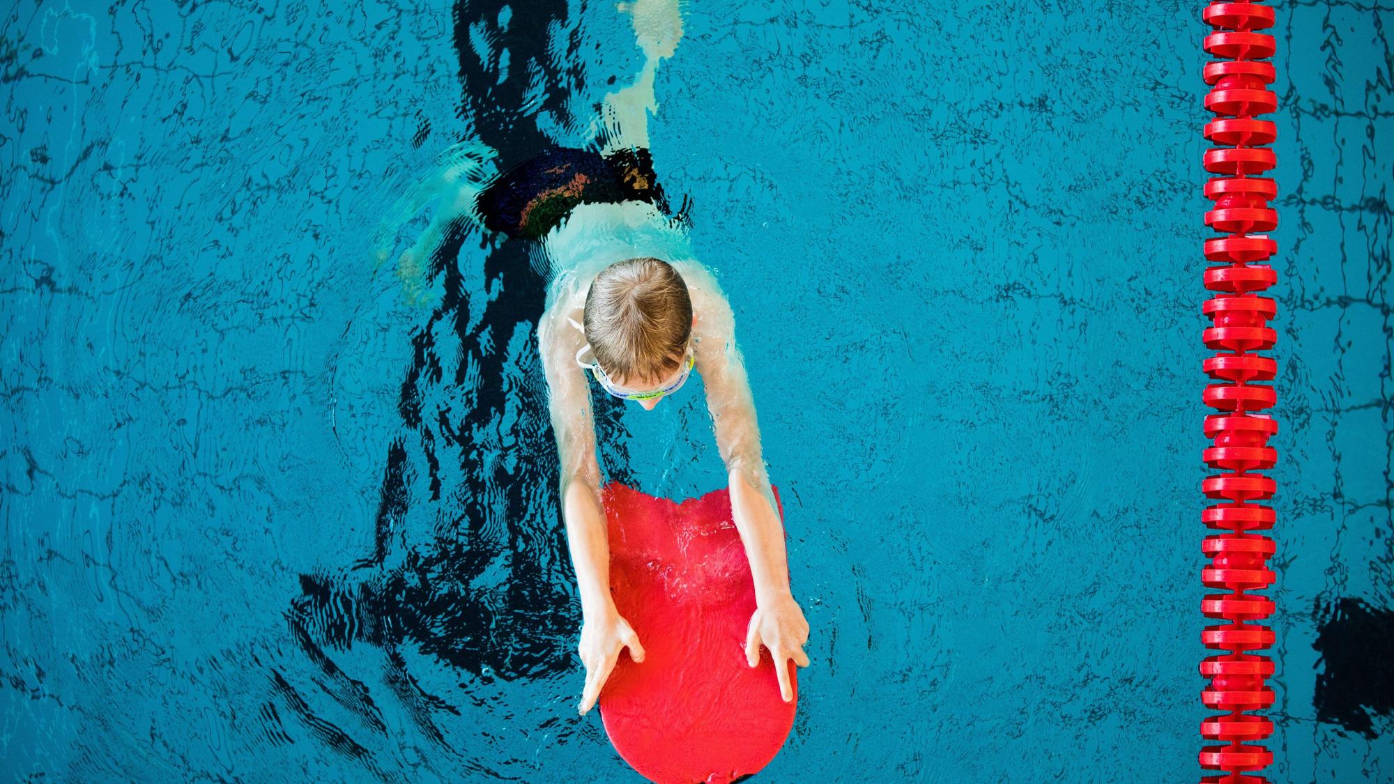 Ein Junge nimmt an einem Schwimmkurs für Kinder teil. Er hält sich mit einem roten Schwimmbrett über Wasser.