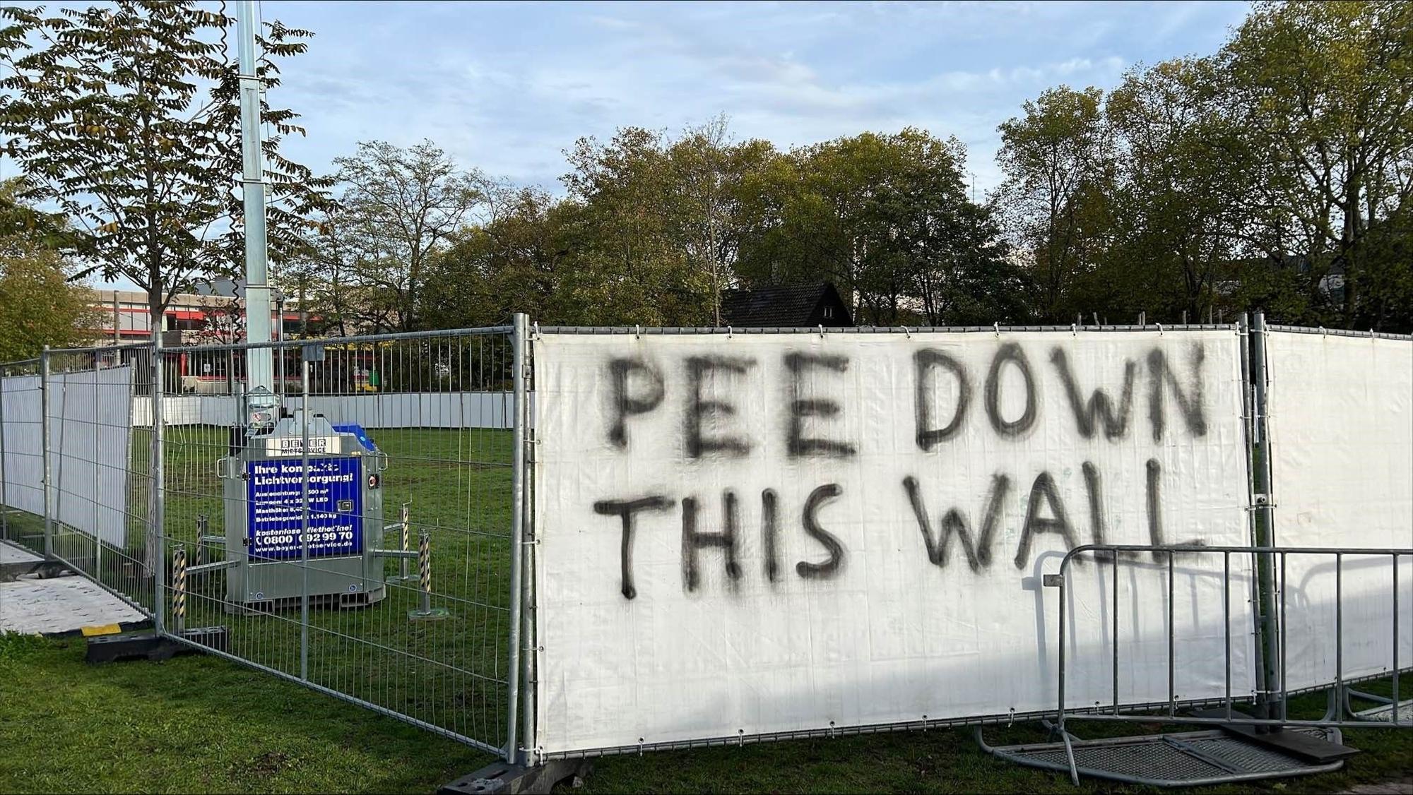 Auf einem Transparent, welches an einem Bauzaun vor den Uniwiesen hängt, steht „Pee Down This Wall“