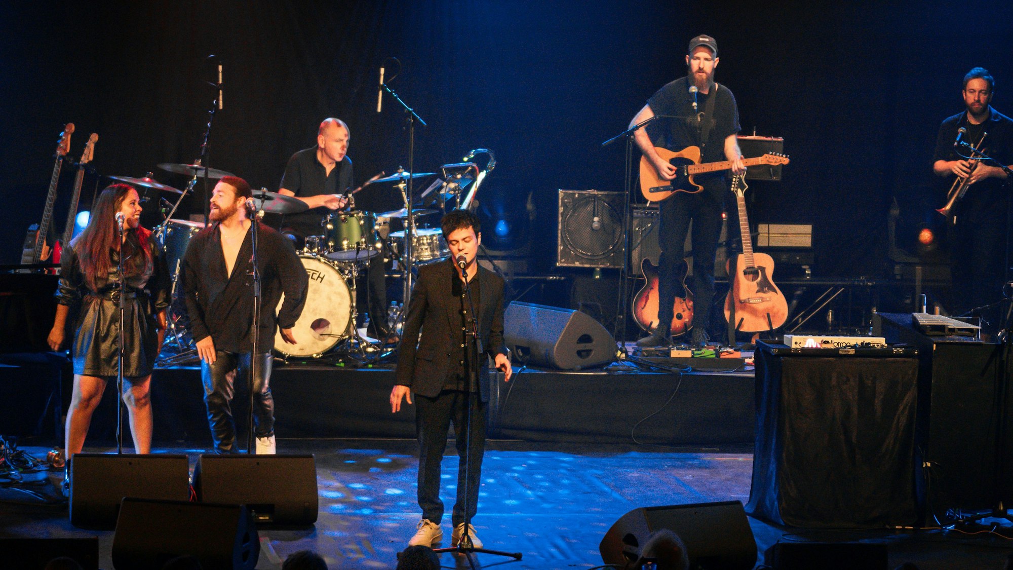 Auf der Bühne im Leverkusener Forum steht Jamie Cullum vorne in der Mitte und singt. Links stehen eine Sängerin und ein Sänger am Mikro und tanzen. Hinter Culum ist ein Schlagzeuger, ein Gitarrist und ein Mann mit Trompete, die ihre Instrumente spielen.