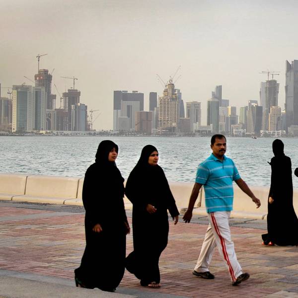 Zwei katarische Frauen und ein Mann spazieren vor der Skyline von Doha. Im Hintergrund spazieren drei Frauen. Alle Frauen tragen lange, schwarze Kleider und Kopftücher. Der Mann trägt eine weiße Hose und ein hellblaues Hemd.