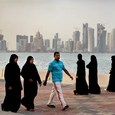 Zwei katarische Frauen und ein Mann spazieren vor der Skyline von Doha. Im Hintergrund spazieren drei Frauen. Alle Frauen tragen lange, schwarze Kleider und Kopftücher. Der Mann trägt eine weiße Hose und ein hellblaues Hemd.