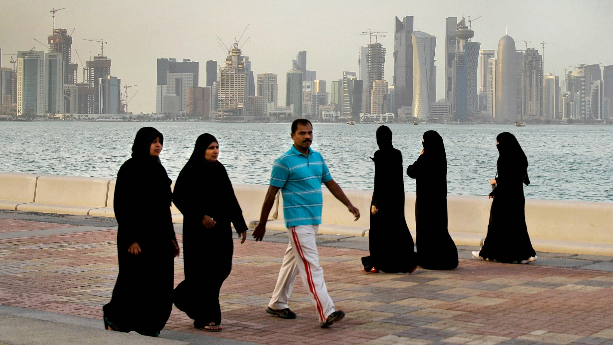 Zwei katarische Frauen und ein Mann spazieren vor der Skyline von Doha. Im Hintergrund spazieren drei Frauen. Alle Frauen tragen lange, schwarze Kleider und Kopftücher. Der Mann trägt eine weiße Hose und ein hellblaues Hemd.