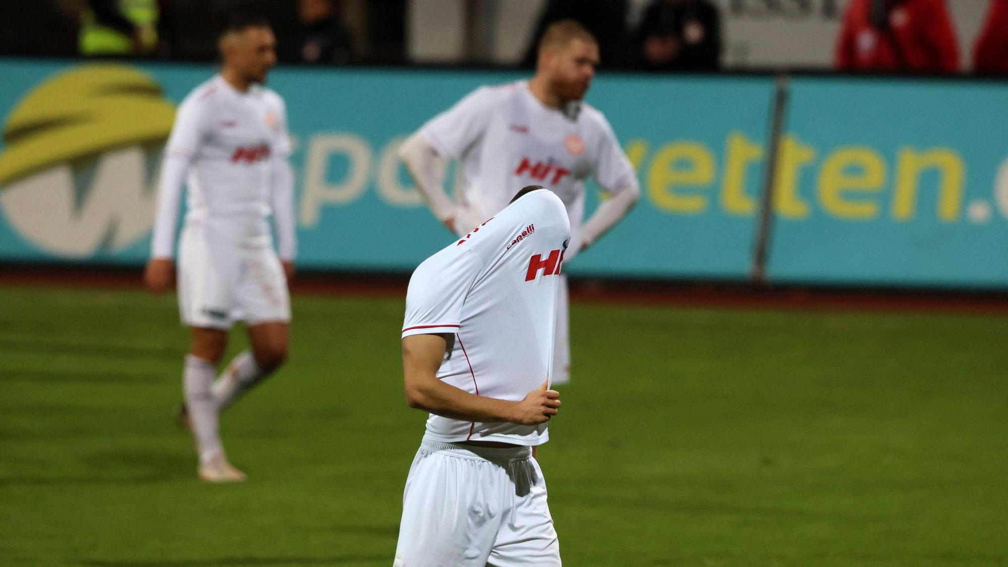 Drei Fußballspieler gehen vom Platz. Der vordere zieht sich enttäuscht das Trikot über den Kopf.