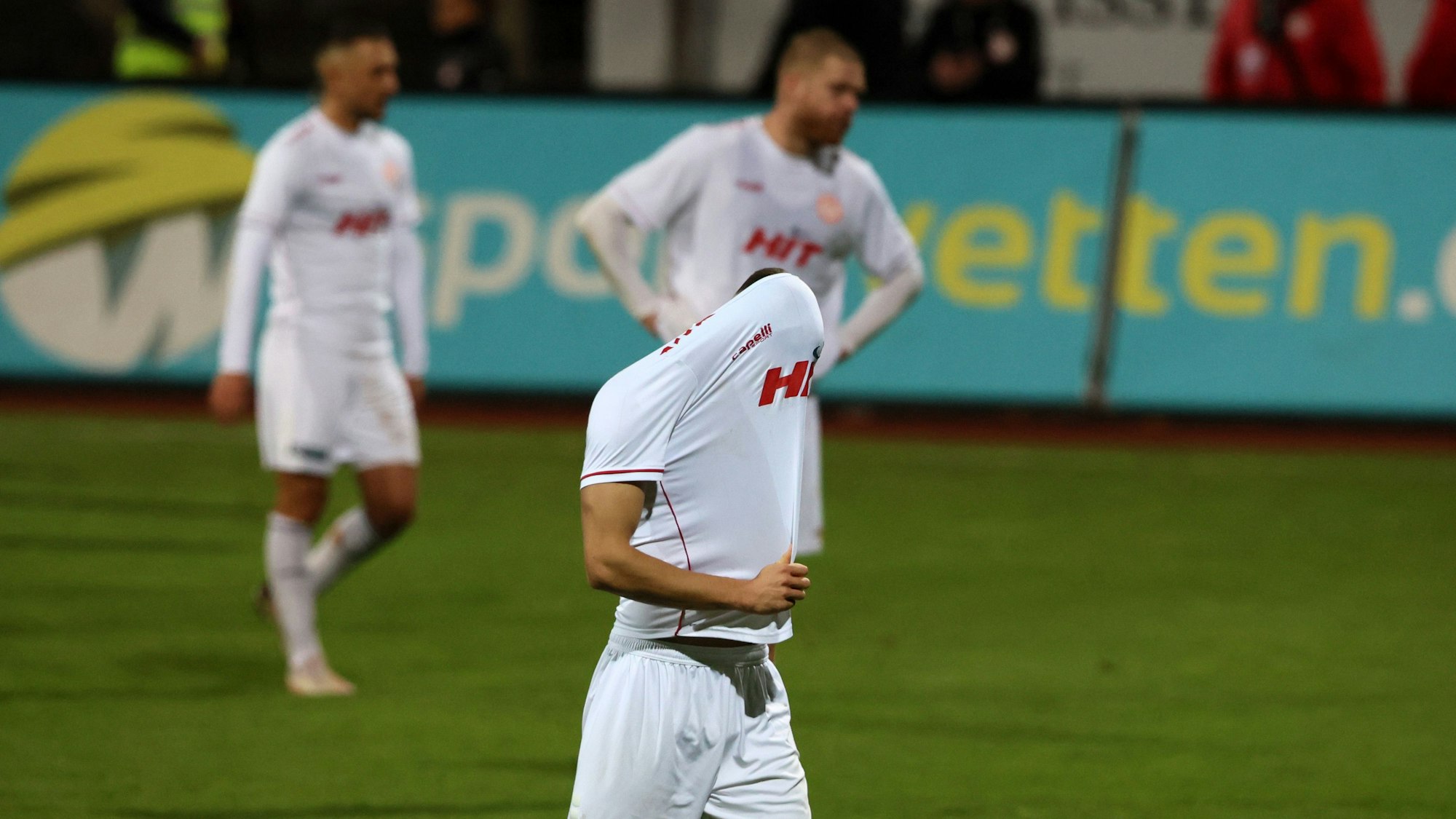Drei Fußballspieler gehen vom Platz. Der vordere zieht sich enttäuscht das Trikot über den Kopf.
