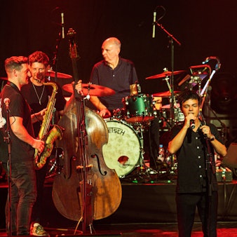 Jamie Cullum steht vorne auf der Bühne und singt. Im Hintergrund sind Musiker mit Saxofon, Kontrabass, Schlagzeug und Gitarre.