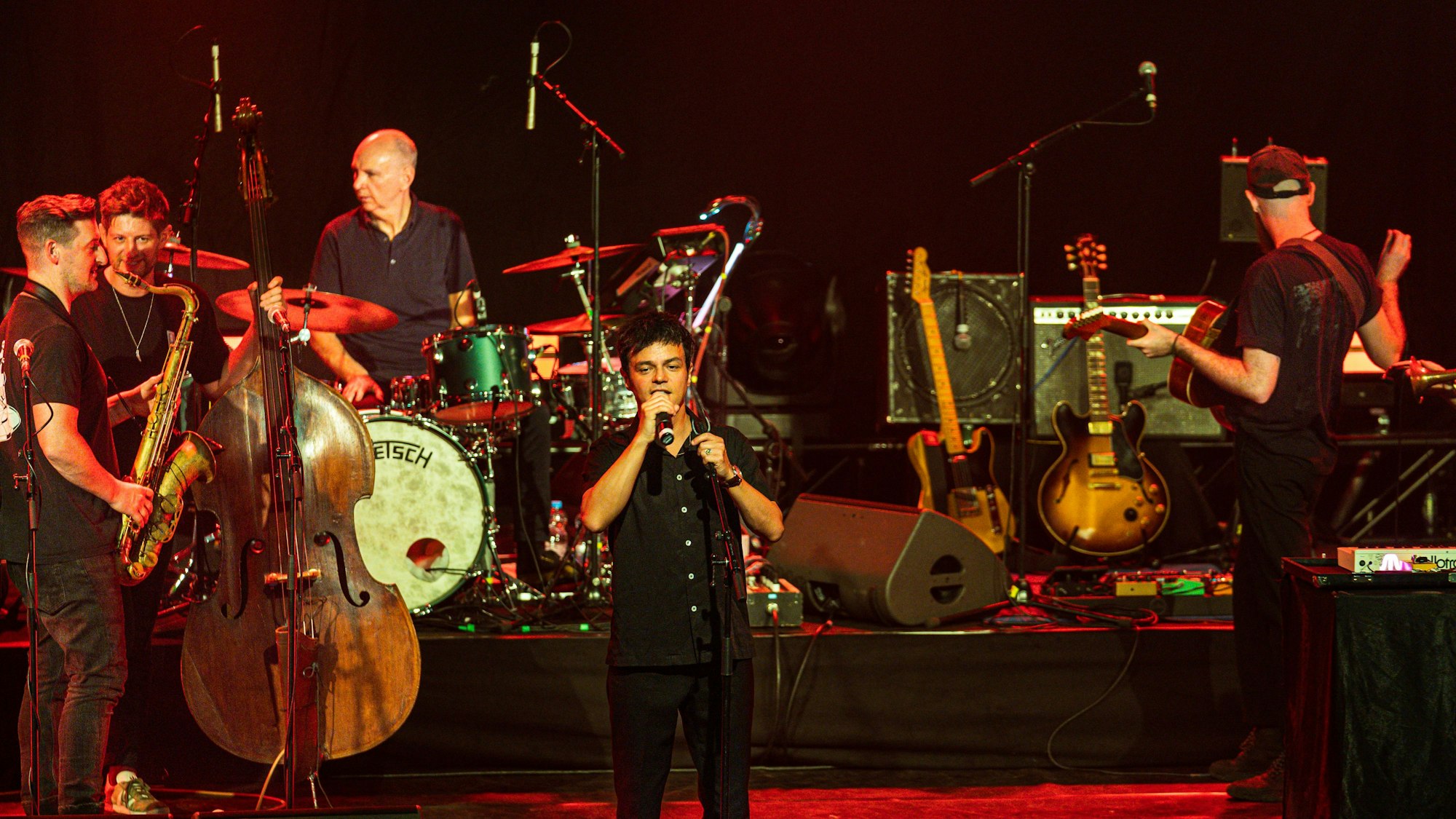 Jamie Cullum steht vorne auf der Bühne und singt. Im Hintergrund sind Musiker mit Saxofon, Kontrabass, Schlagzeug und Gitarre.