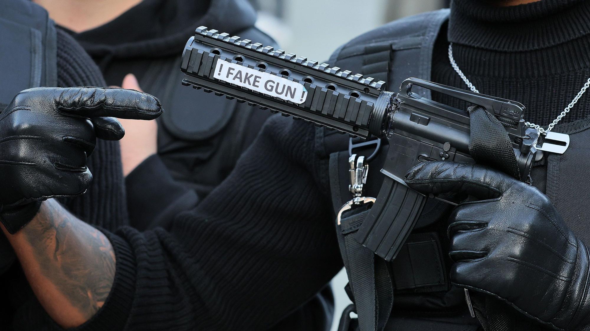 Ein Kostumierter trägt eine falsche Waffe mit der Aufschrift „fake gun“ vor sich.