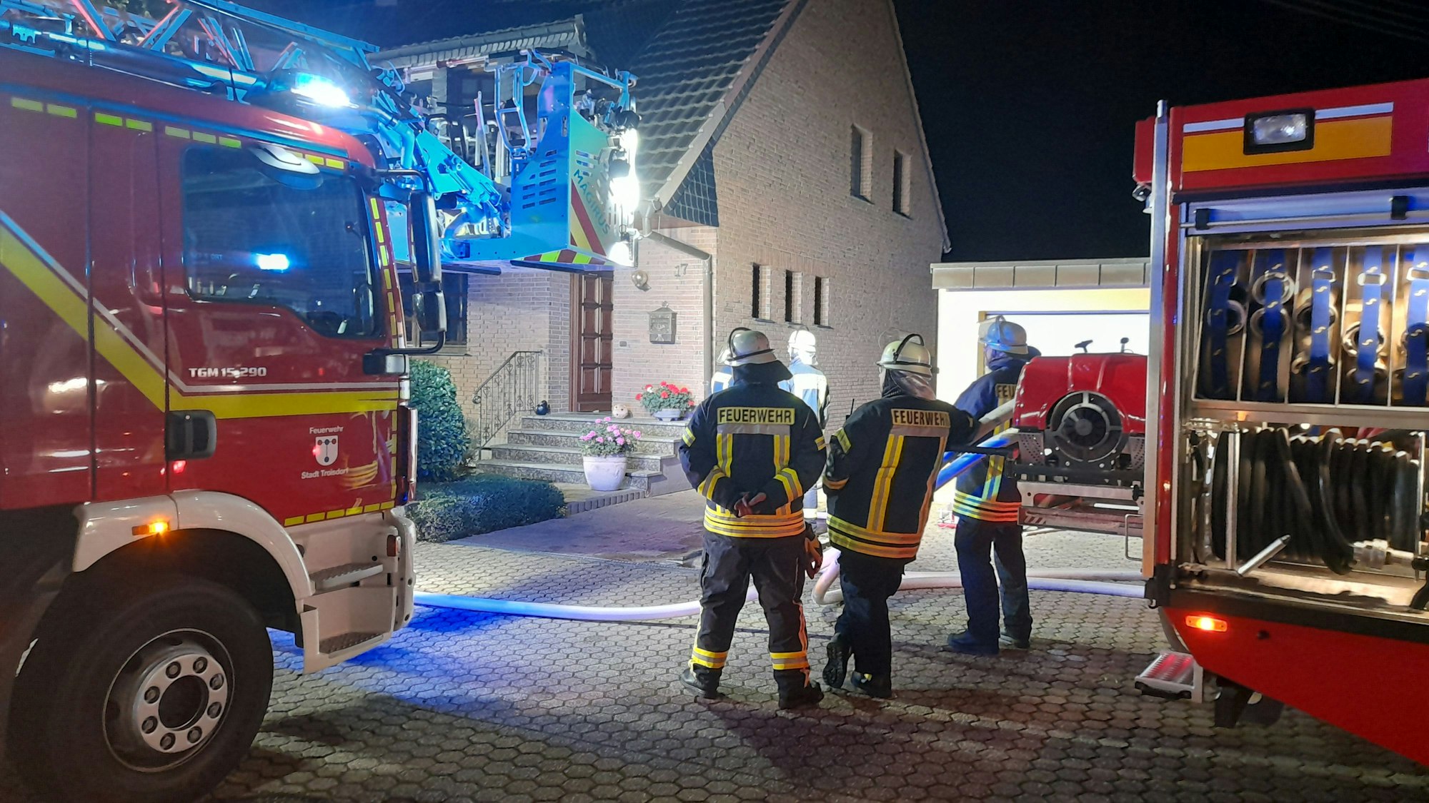 Feuerwehrleute stehen vor einem Haus. Zwei Feuerwehrautos stehen neben ihnen.