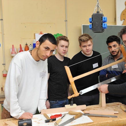 Ausbilder Arnulf Rückamp steht mit seinen Lehrlingen um einen Tisch mit Werkzeug und sägt Holz.