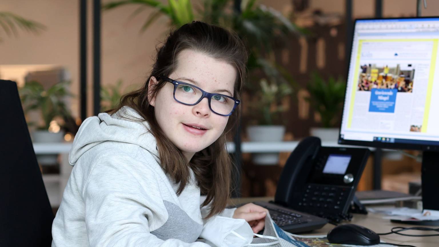 Die gehandicapte Schülerin Aimée Heimbach sitzt an einem Schreibtisch in der Lokalredaktion des Kölner Stadt-Anzeigers vor einem großen Monitor, auf dem die aktuelle Zeitungsseite zu sehen ist.