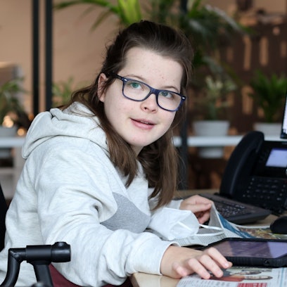 Die gehandicapte Schülerin Aimée Heimbach sitzt an einem Schreibtisch in der Lokalredaktion des Kölner Stadt-Anzeigers vor einem großen Monitor, auf dem die aktuelle Zeitungsseite zu sehen ist.