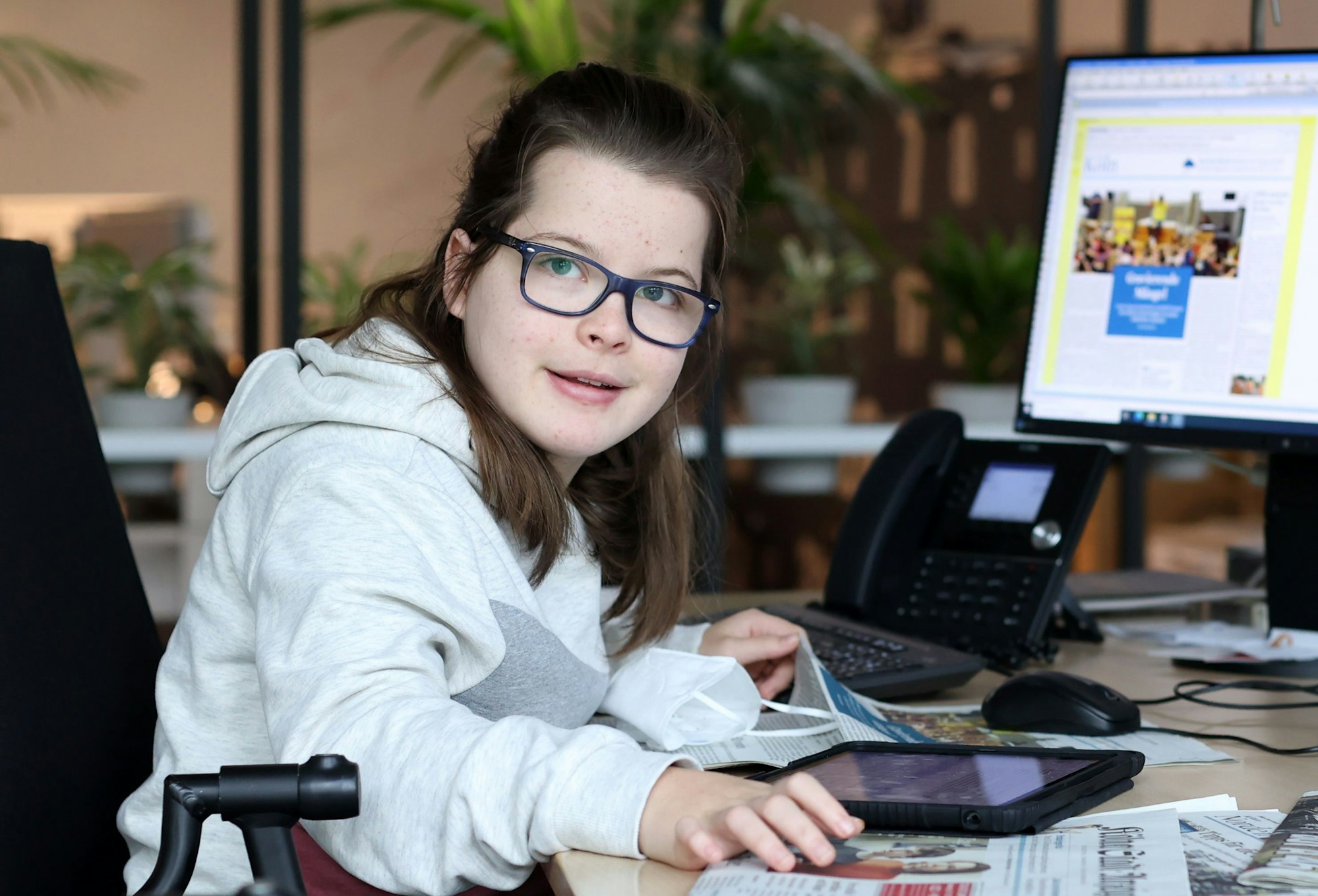 Die gehandicapte Schülerin Aimée Heimbach sitzt an einem Schreibtisch in der Lokalredaktion des Kölner Stadt-Anzeigers vor einem großen Monitor, auf dem die aktuelle Zeitungsseite zu sehen ist.