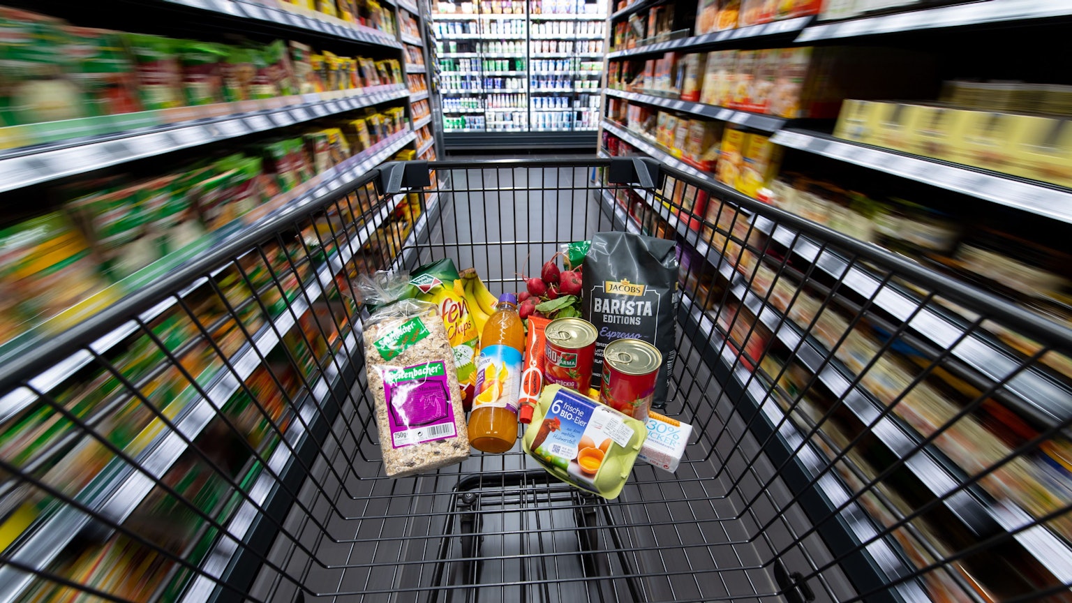 Ein Einkaufswagen, gefüllt mit Konserven, Eiern und einigen anderen Lebensmitteln, wird durch einen Gang im Supermarkt geschoben.