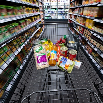 Ein Einkaufswagen, gefüllt mit Konserven, Eiern und einigen anderen Lebensmitteln, wird durch einen Gang im Supermarkt geschoben.