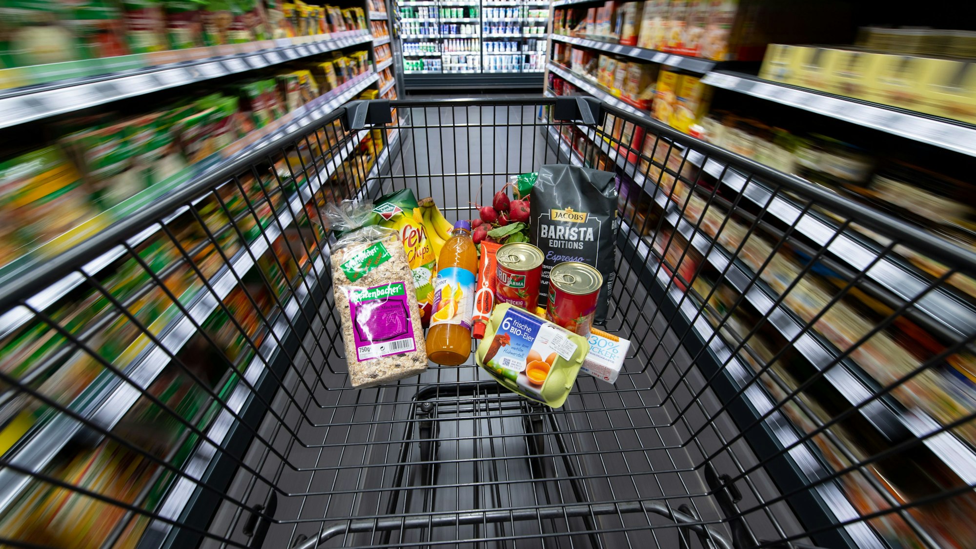 Ein Einkaufswagen, gefüllt mit Konserven, Eiern und einigen anderen Lebensmitteln, wird durch einen Gang im Supermarkt geschoben.