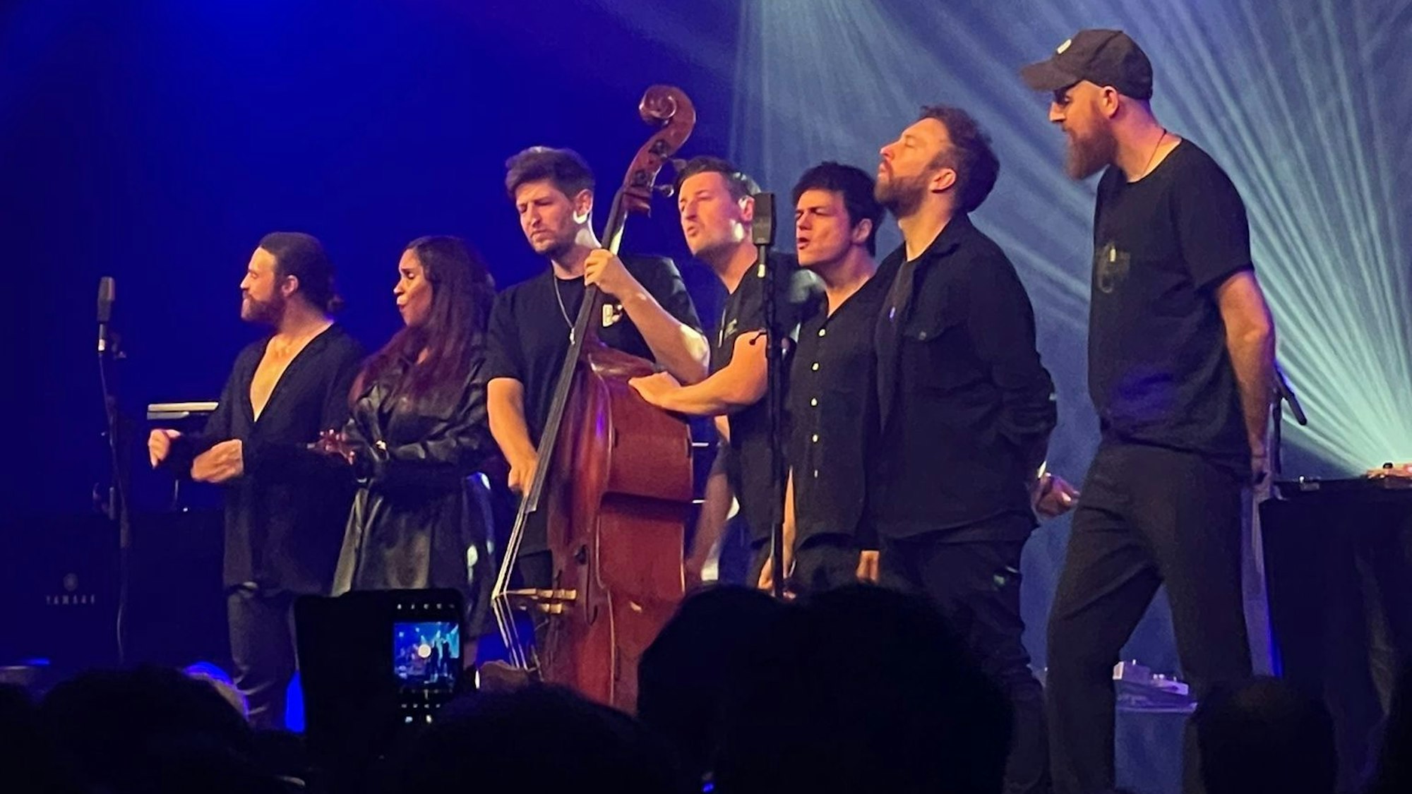 Auf der Bühne stehen sieben Musiker und Musikerinnen. Es ist die Band um Jamie Cullum. In der Mitte steht ein Kontrabass, das einer der Band-Mitglieder spielt. Ein andere benutzt es als Trommel. Alle singen