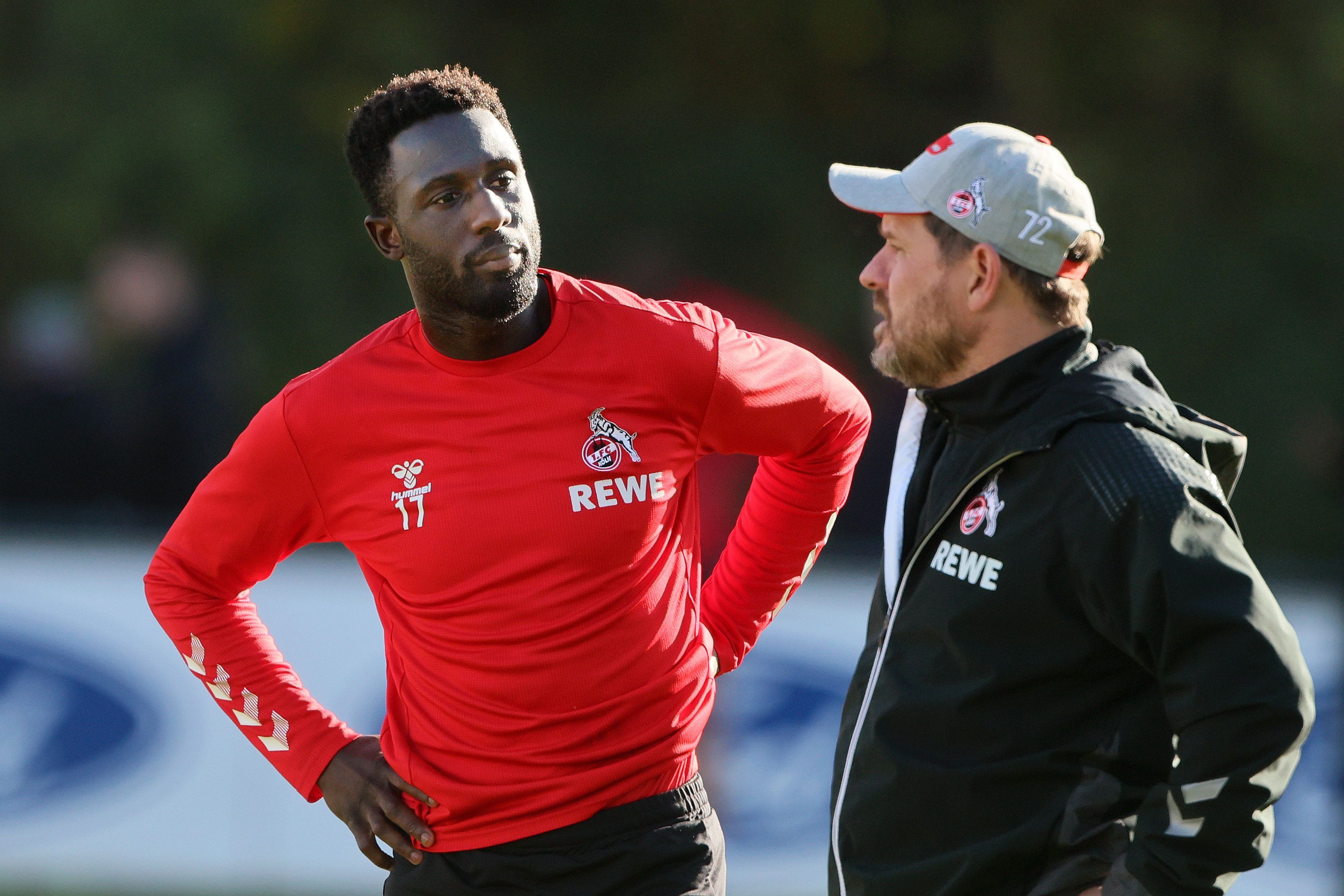 Steffen Baumgart in schwarzer Trainingsjacke spricht auf dem Platz mit Kingsley Schindler, der die Arme in die Hüfte gestemmt hat und ein rotes Trainingsshirt mit langen Ärmeln trägt.