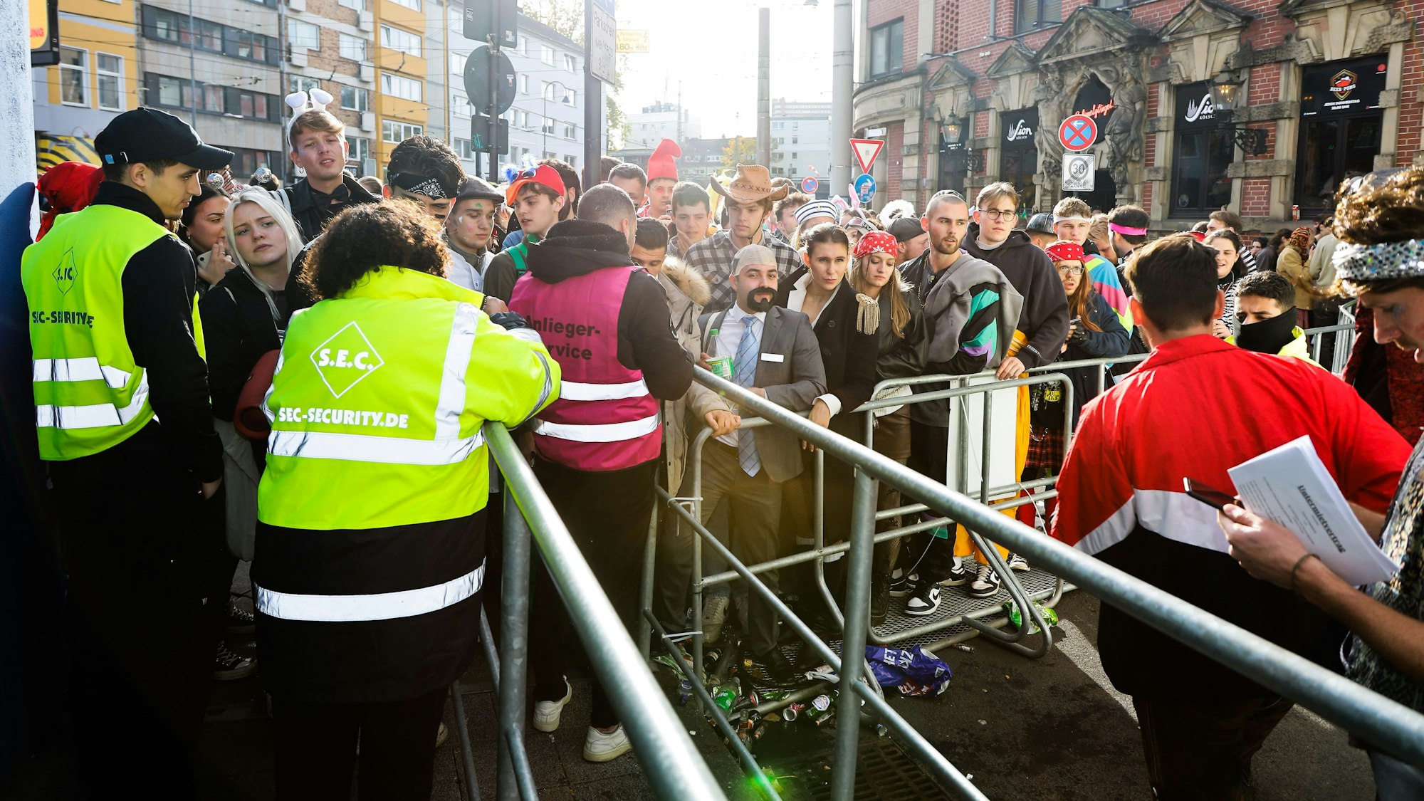 Zwei Ordner stehen am 11.11. vor einer Absperrung im Zülpicher Viertel, vor der sich kostümierte Menschen drängen.