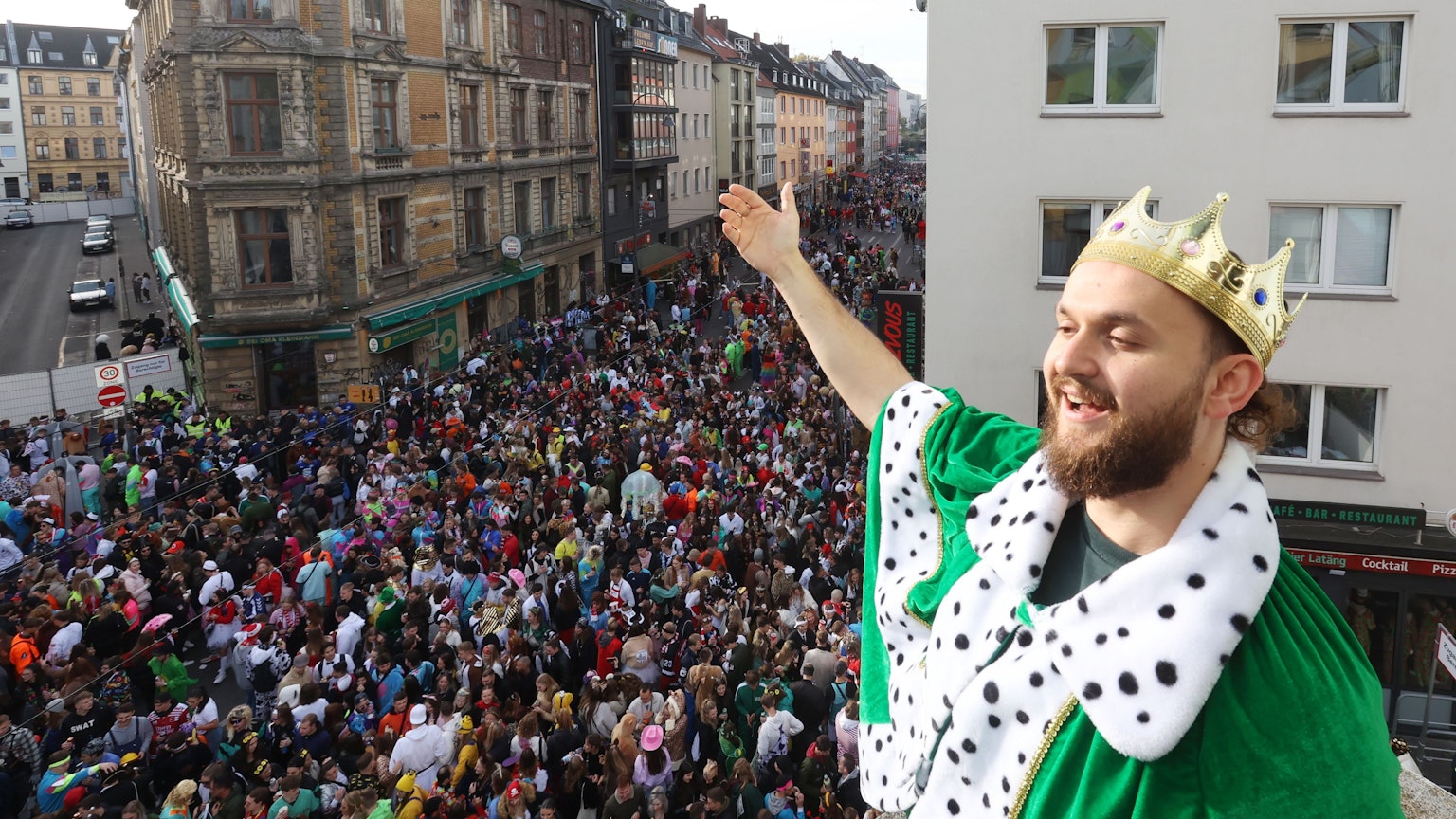 Ein junger Mann steht mit einer Krone auf dem Balkon. Unter ihm die volle Zülpicher Straße.