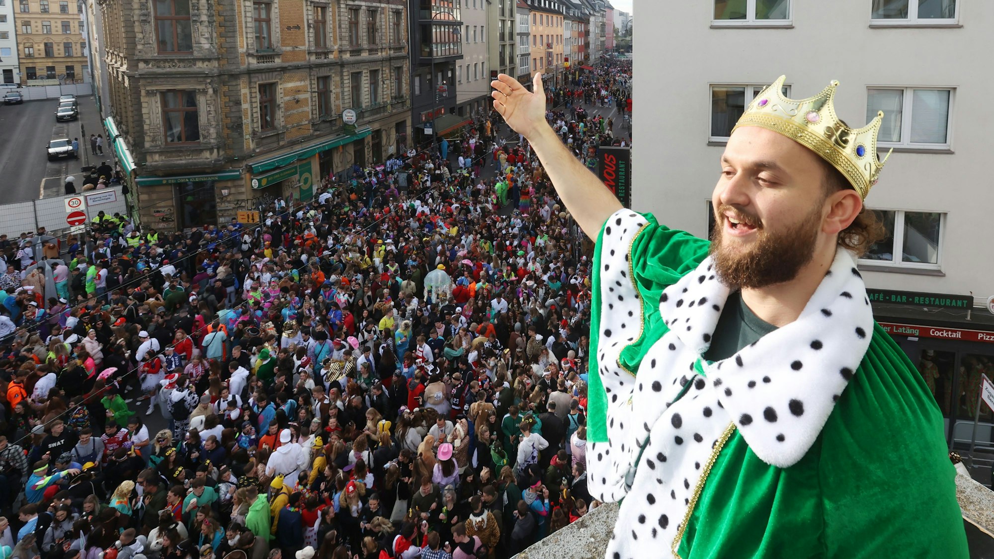 Ein junger Mann steht mit einer Krone auf dem Balkon. Unter ihm die volle Zülpicher Straße.