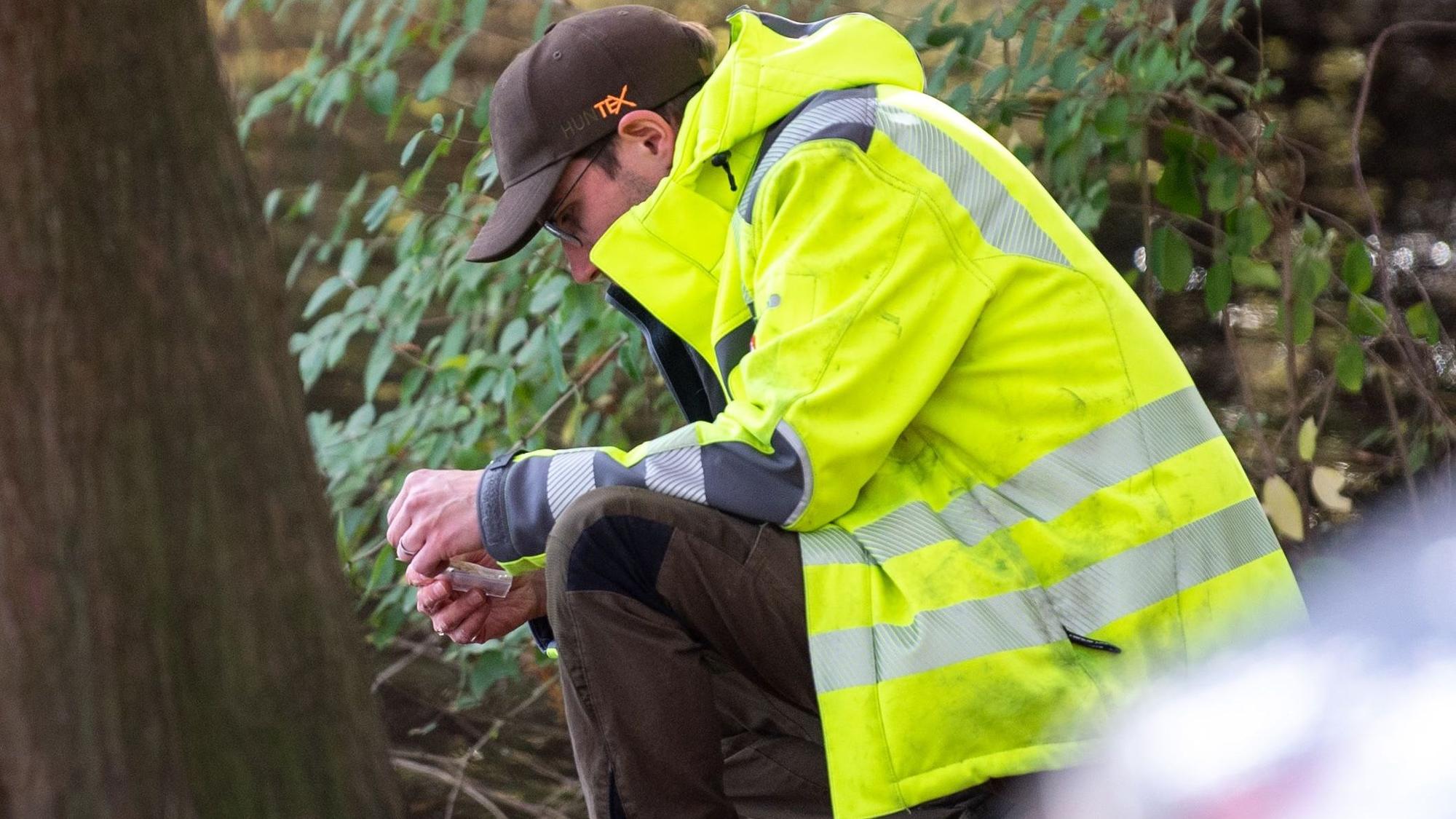 Ein Mitarbeiter der Unteren Wasserbehörde kniet auf dem Boden und entnimmt eine Wasserprobe. Er trägt eine gelbe Jacke.