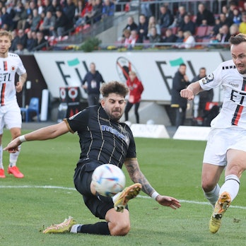 Ein Fußballspieler flankt den Ball vors Tor und sein Gegenspieler versucht es zu verhindern.