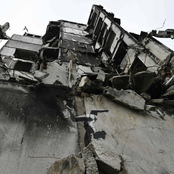 Ein vom Krieg zerstörtes Haus in der Ukraine ist zu sehen, im Vordergrund ist auf einer Betonmauer eine gemalte Tänzerin zu sehen, die einen Handstand macht.