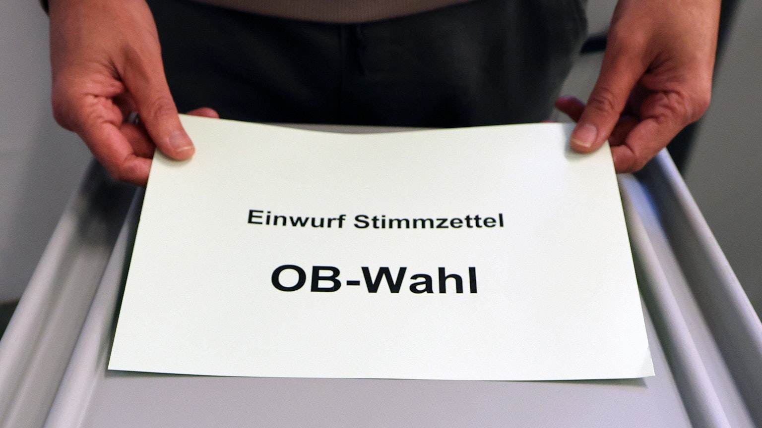 Ein Zettel mit der Aufschrift „Einfwurf Stimmzettel OB-Wahl“ wird vor einer grauen Wahlurne gezeigt.