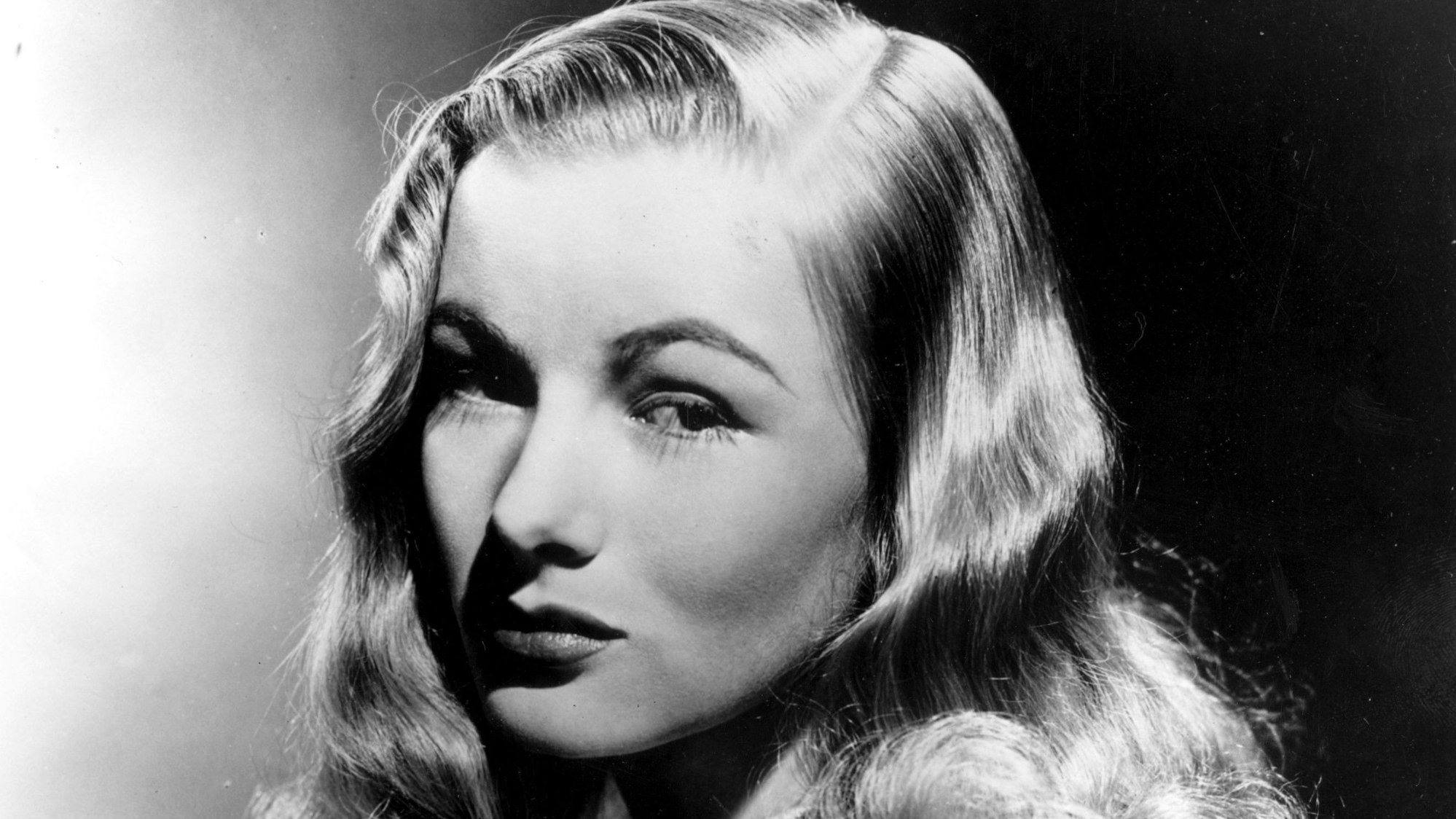 Die Schauspielerin Veronica Lake in Schwarzweiß. Ihr Markenzeichen, eine Haarlocke, die auf einer Seite über ihr Gesicht hängt.