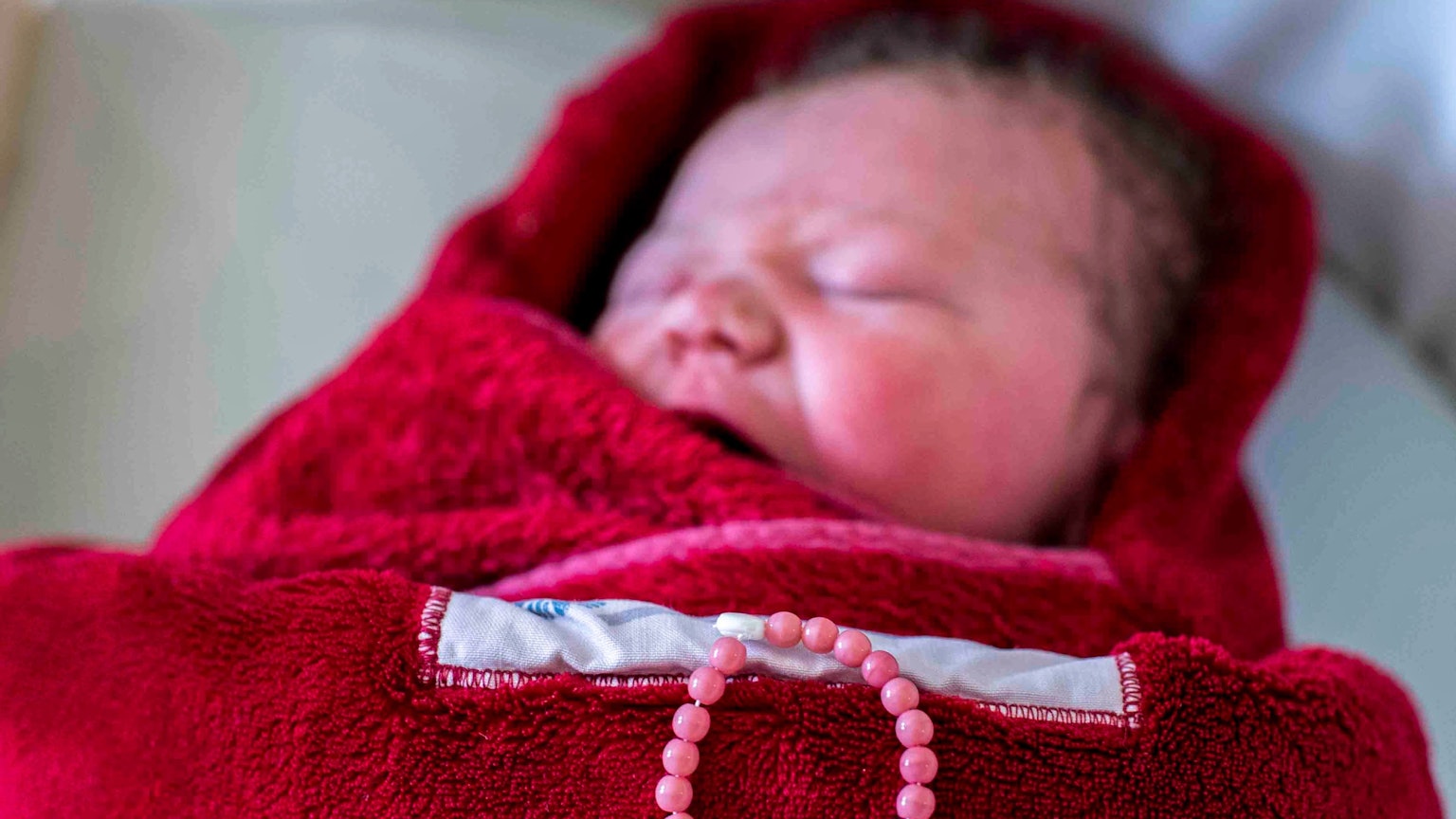 Ein neugeborenes Kind liegt in eine rote Decke eingewickelt in einem Bett. Auf der Decke liegt eine Kette mit dem Namen des Kindes: Juna.