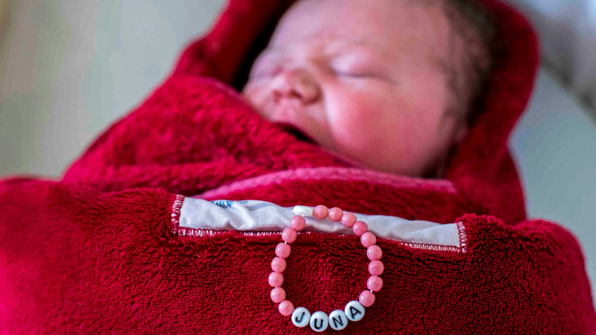 Ein neugeborenes Kind liegt in eine rote Decke eingewickelt in einem Bett. Auf der Decke liegt eine Kette mit dem Namen des Kindes: Juna.