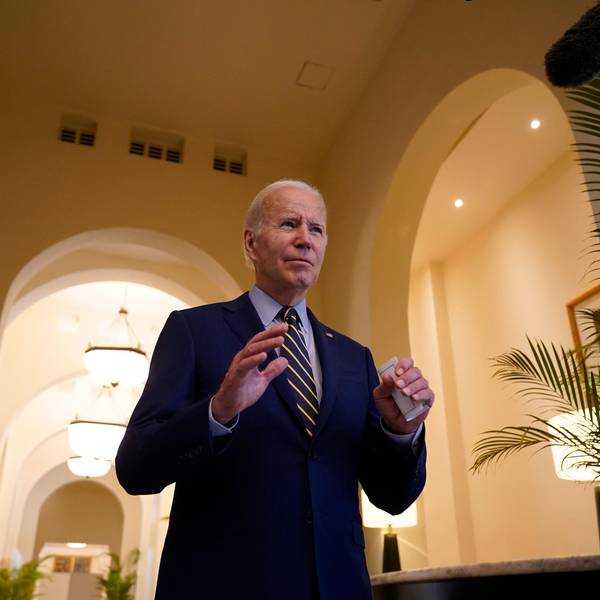 US-Präsident Joe Biden steht in einem blauen Anzug in einem Gebäude mit hohen Torbögen. Er hebt die Hände mit den Handflächen nach unten.