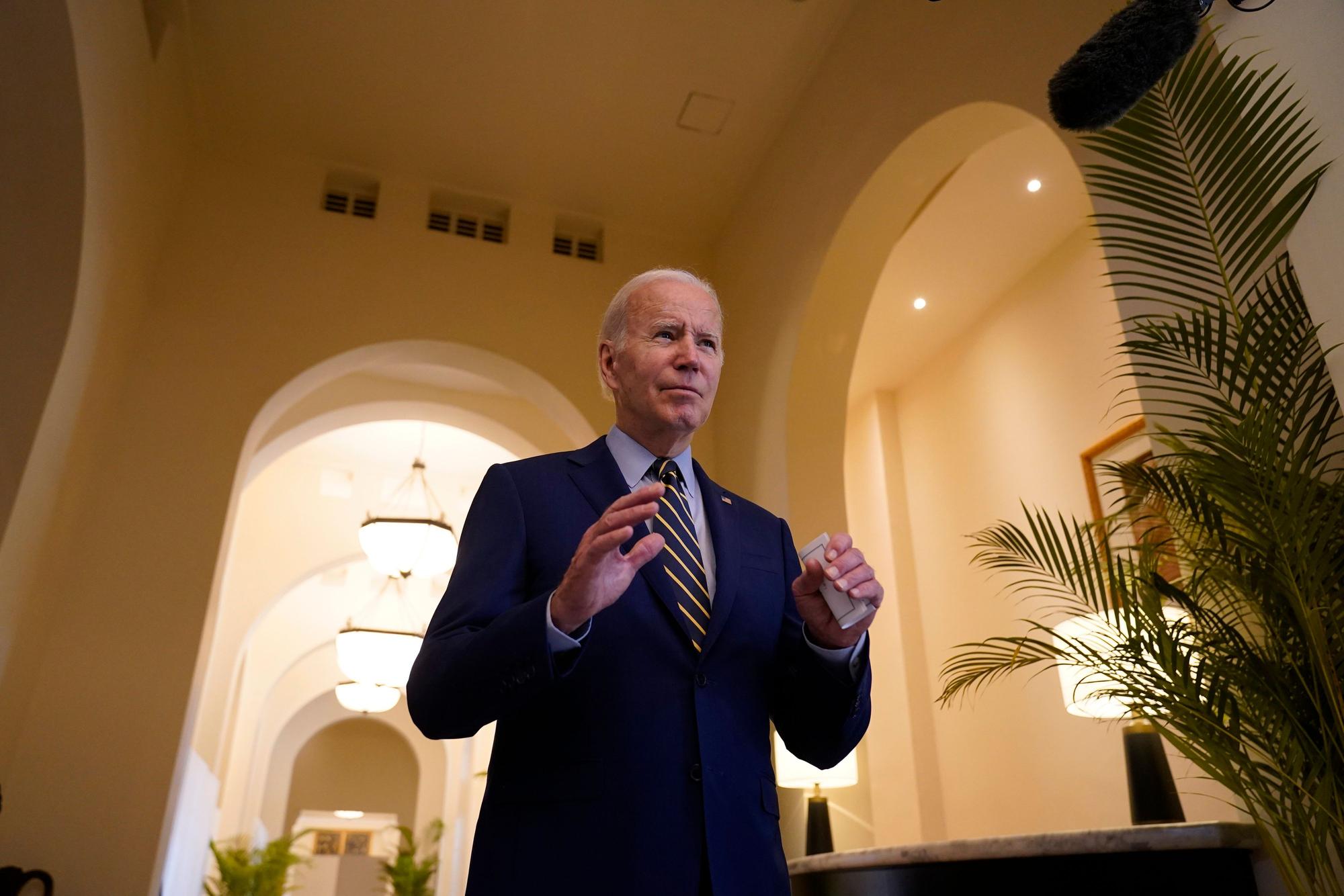 US-Präsident Joe Biden steht in einem blauen Anzug in einem Gebäude mit hohen Torbögen. Er hebt die Hände mit den Handflächen nach unten.