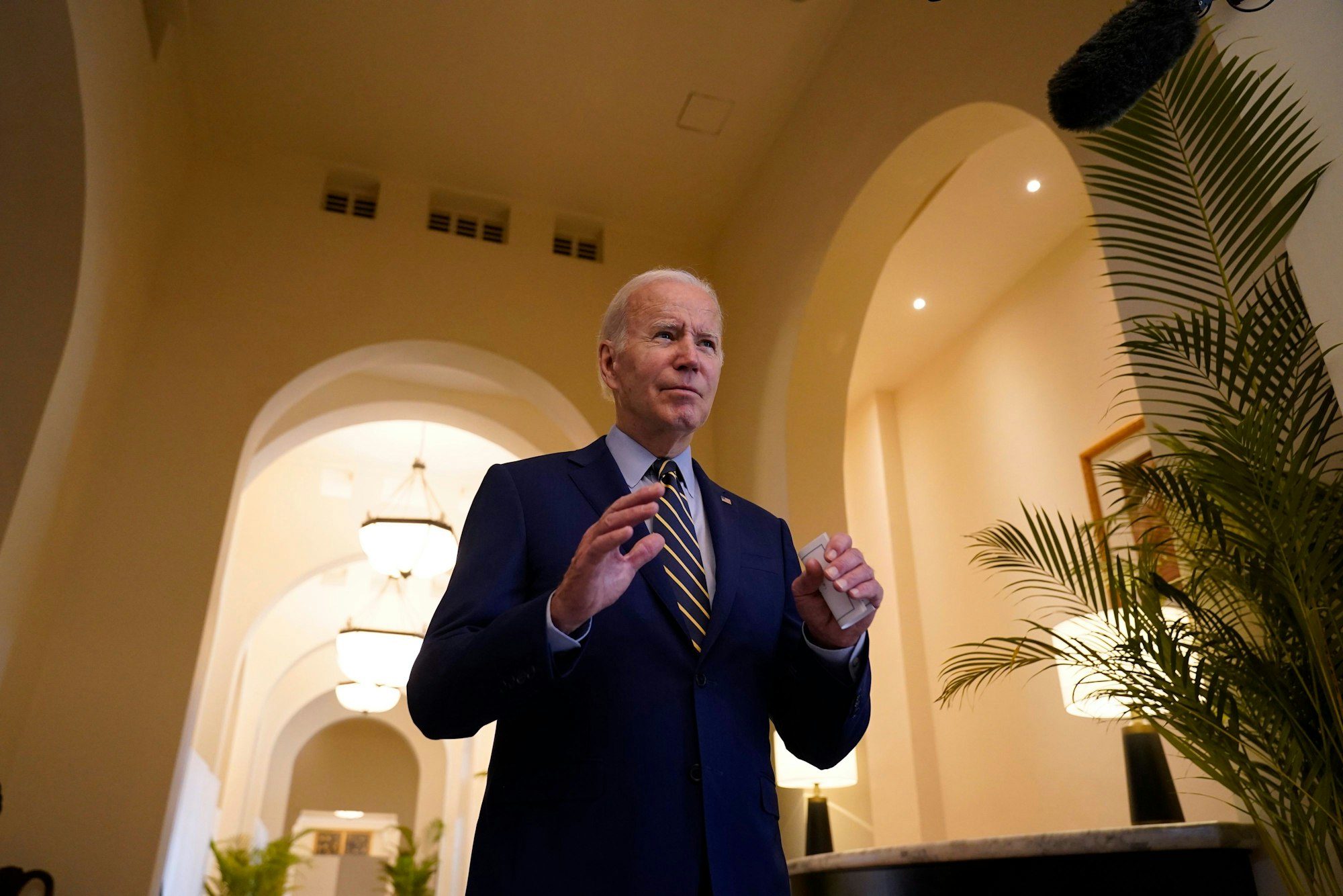 US-Präsident Joe Biden steht in einem blauen Anzug in einem Gebäude mit hohen Torbögen. Er hebt die Hände mit den Handflächen nach unten.