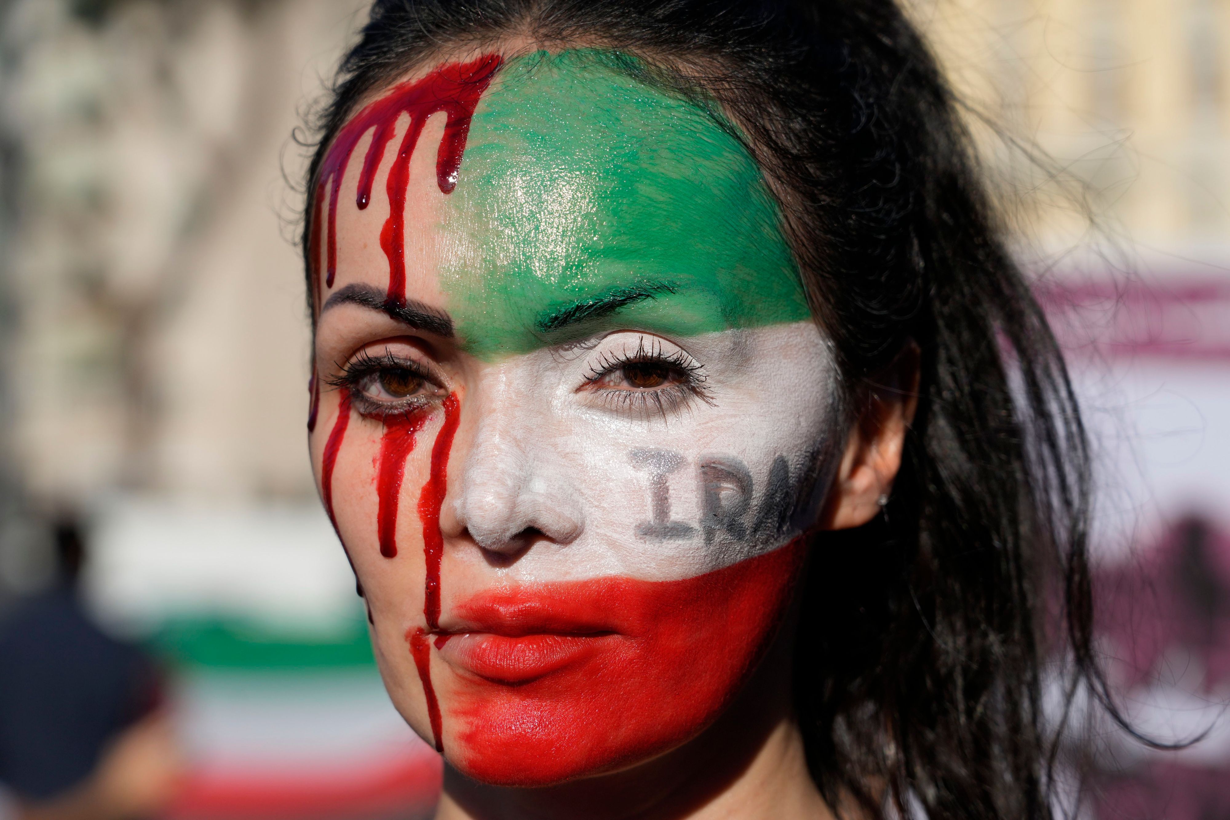 Eine Frau mit bemaltem Gesicht: Auf der linken Hälfte die Iranische Flagge, auf der rechten läuft ihr Kunstblut über die Haut.