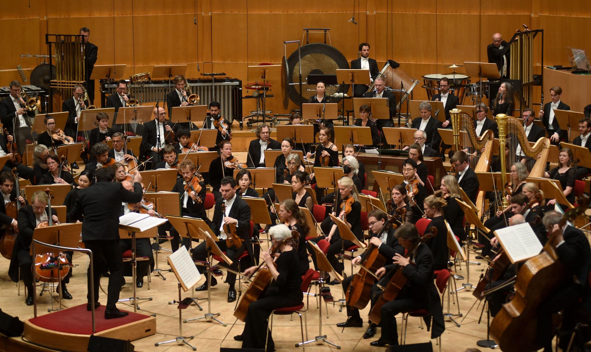 Chef-Dirigent Cristian Macelaru steht mit dem Rücken zur Kamera und dirigiert das WDR-Sinfonieorchester in einem Konzertsaal.